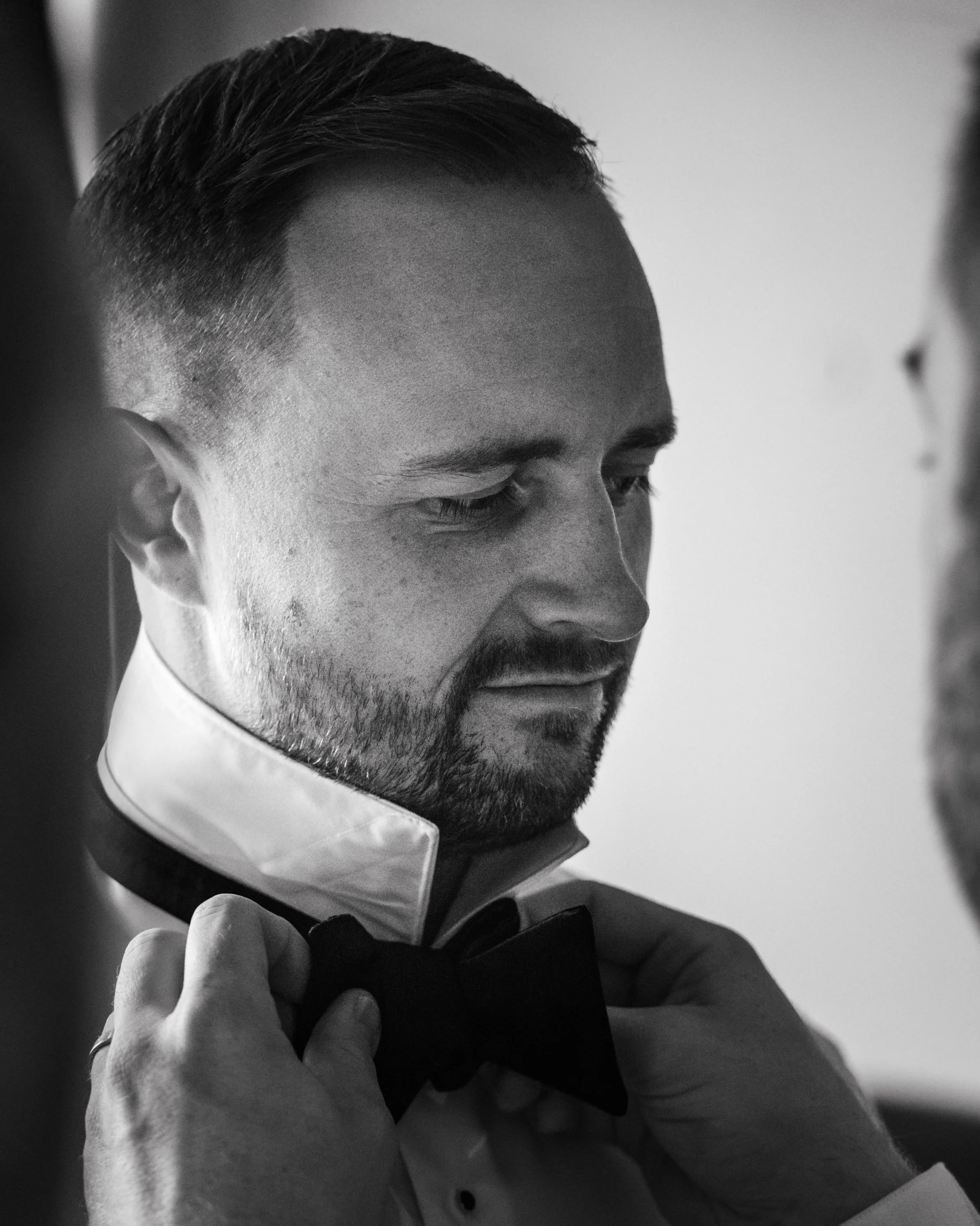 Black and white photo of a man adjusting his bow tie, wearing a formal shirt with the collar up.