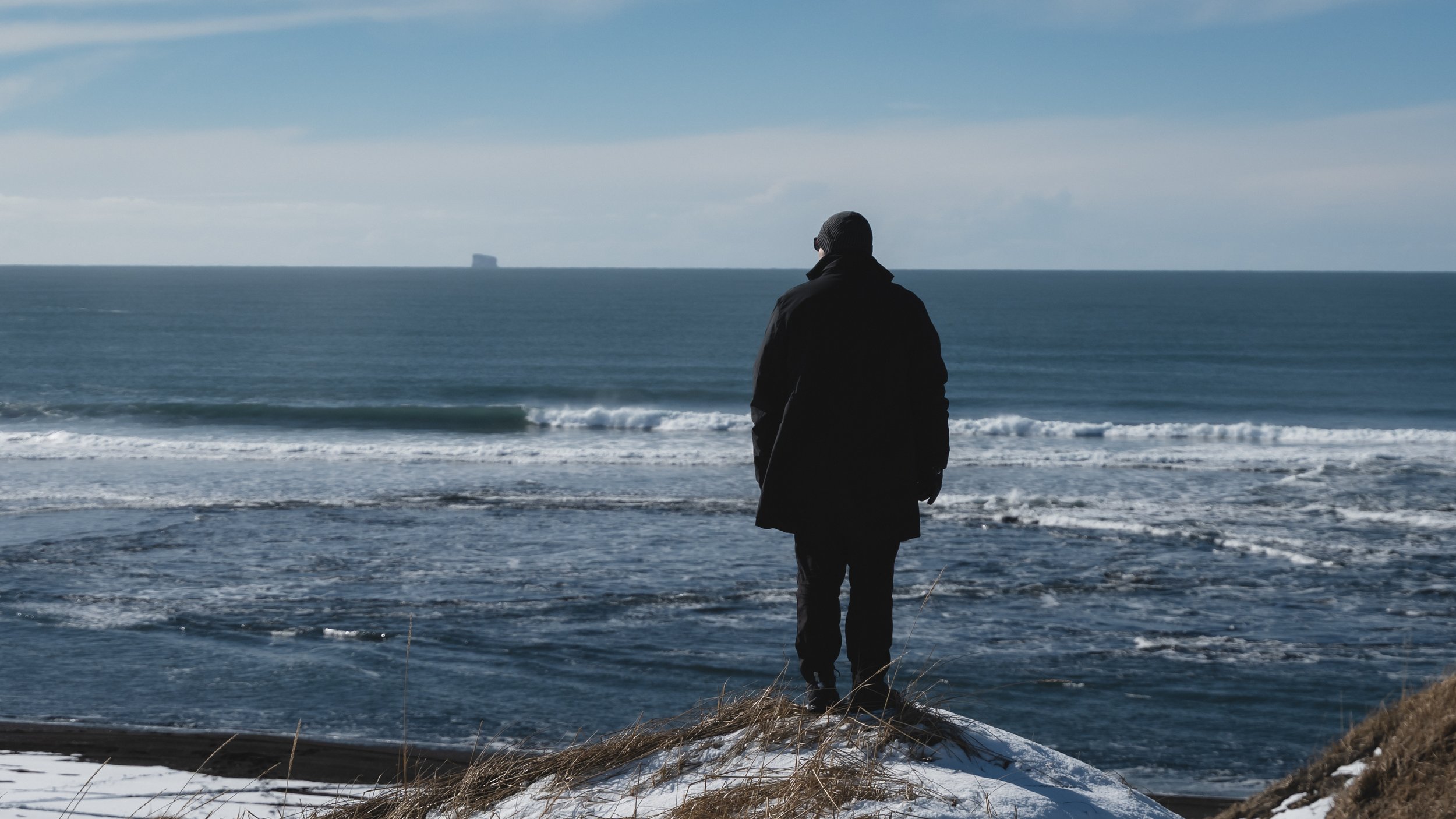 A person dressed in dark clothing standing on a snowy and grassy hill, facing the ocean on a cloudy day.