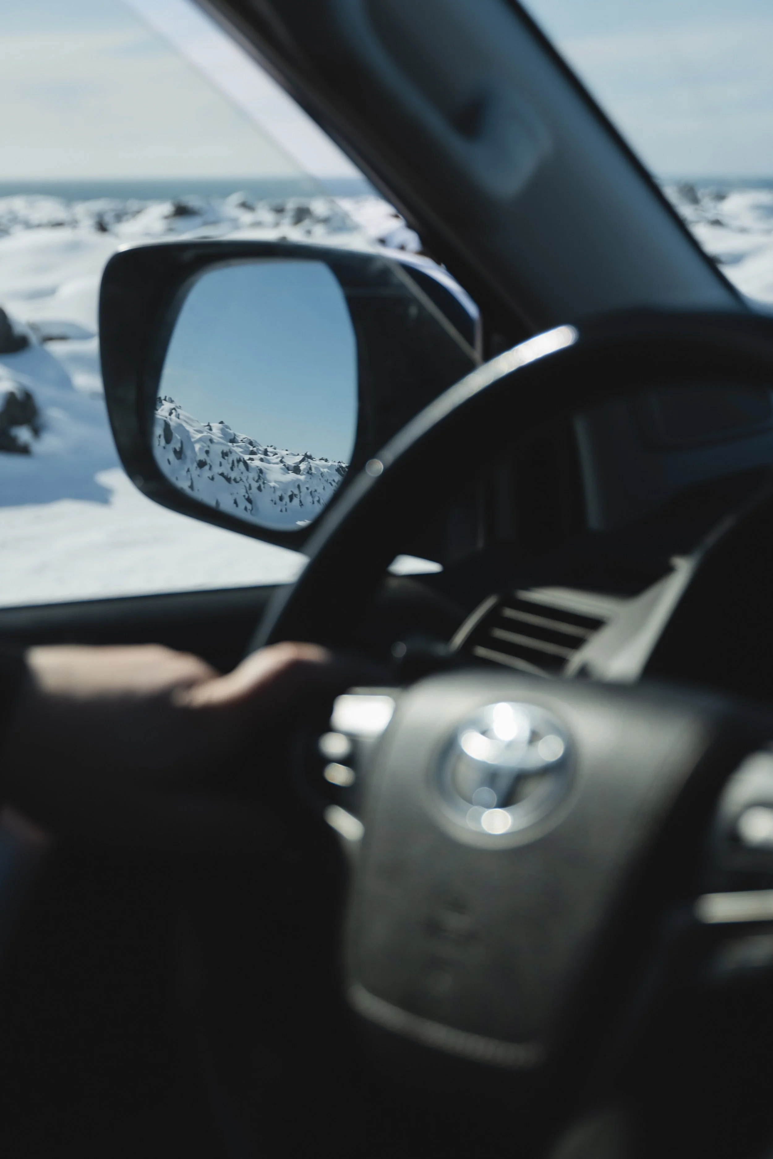 A person driving a vehicle through a snowy landscape, with a view of snow-covered rocks and mountains visible in the side mirror.