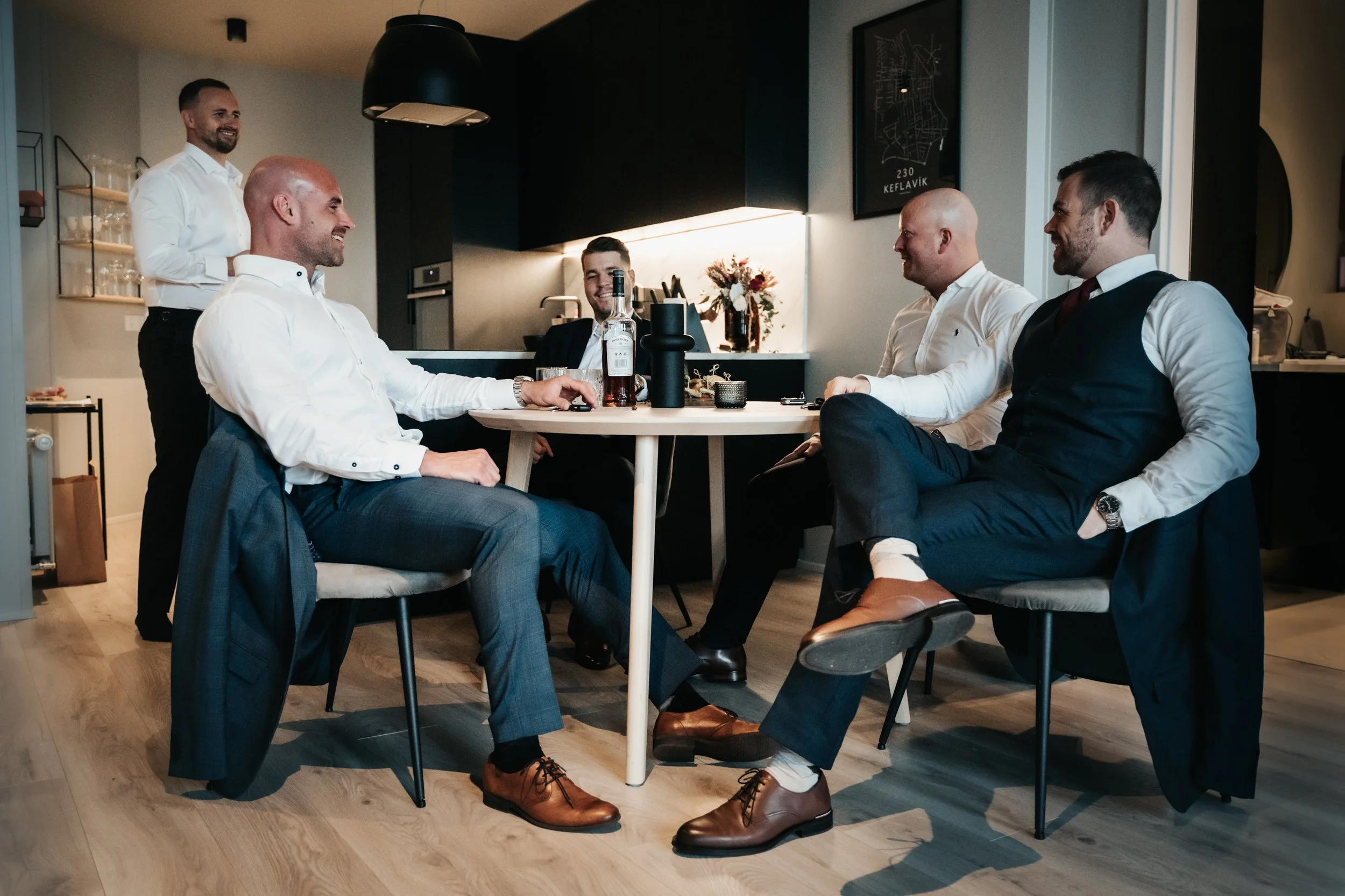 Five men dressed in business attire are gathered around a dining table in a modern kitchen, smiling and engaging in conversation. One man is standing, while the others are seated.