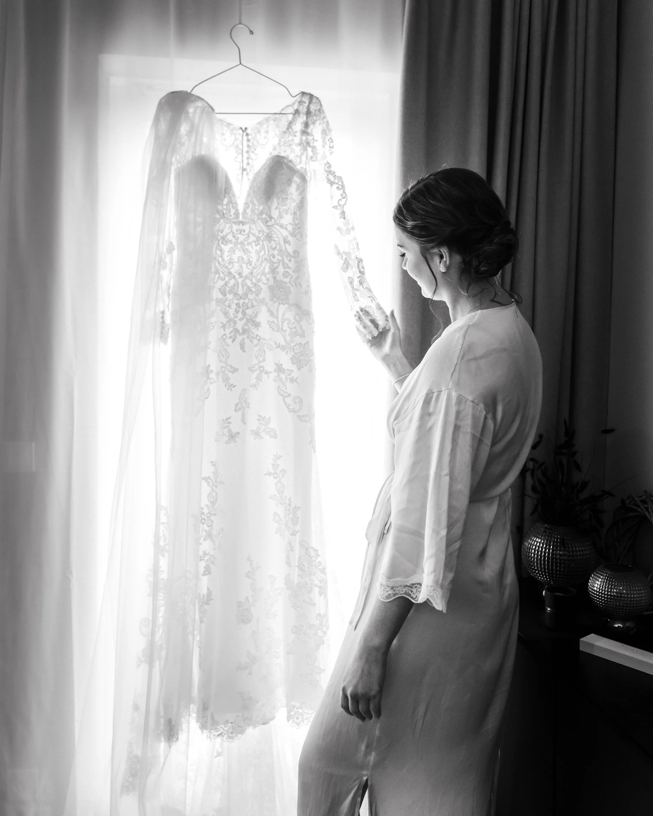 A woman in a satin robe looks at her wedding dress hanging near a window, with curtains and decorative vases in the background.