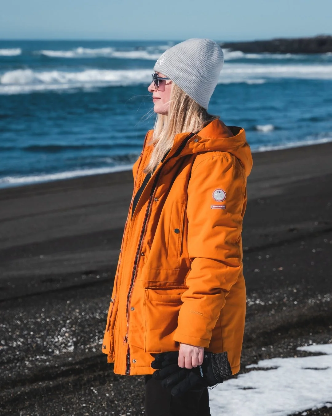 A woman in an orange winter jacket, gray beanie, and sunglasses standing on a black sand beach near the ocean, with waves and a rocky shoreline in the background.