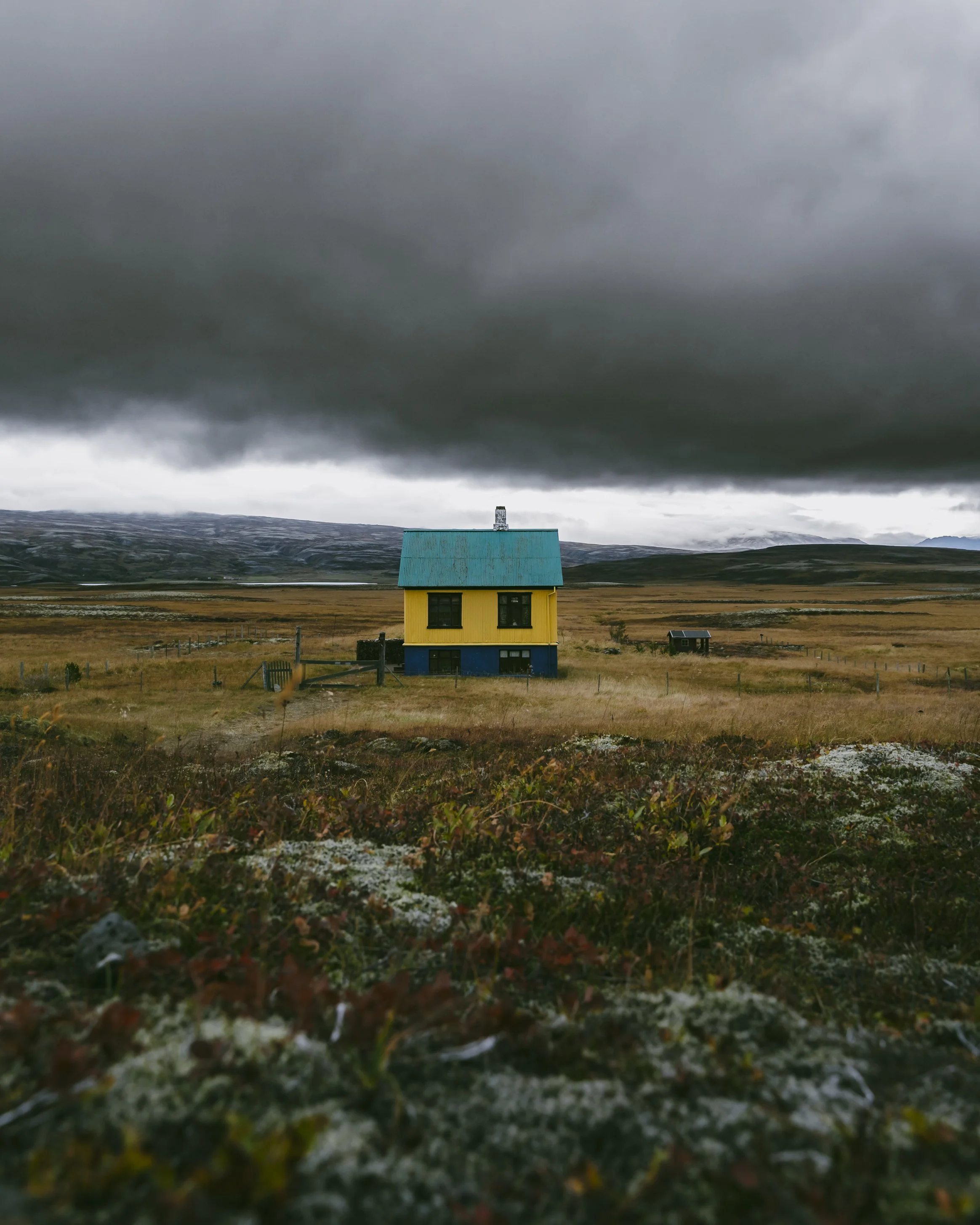 A yellow house with a teal roof stands alone in a vast, open field under dark, cloudy skies.