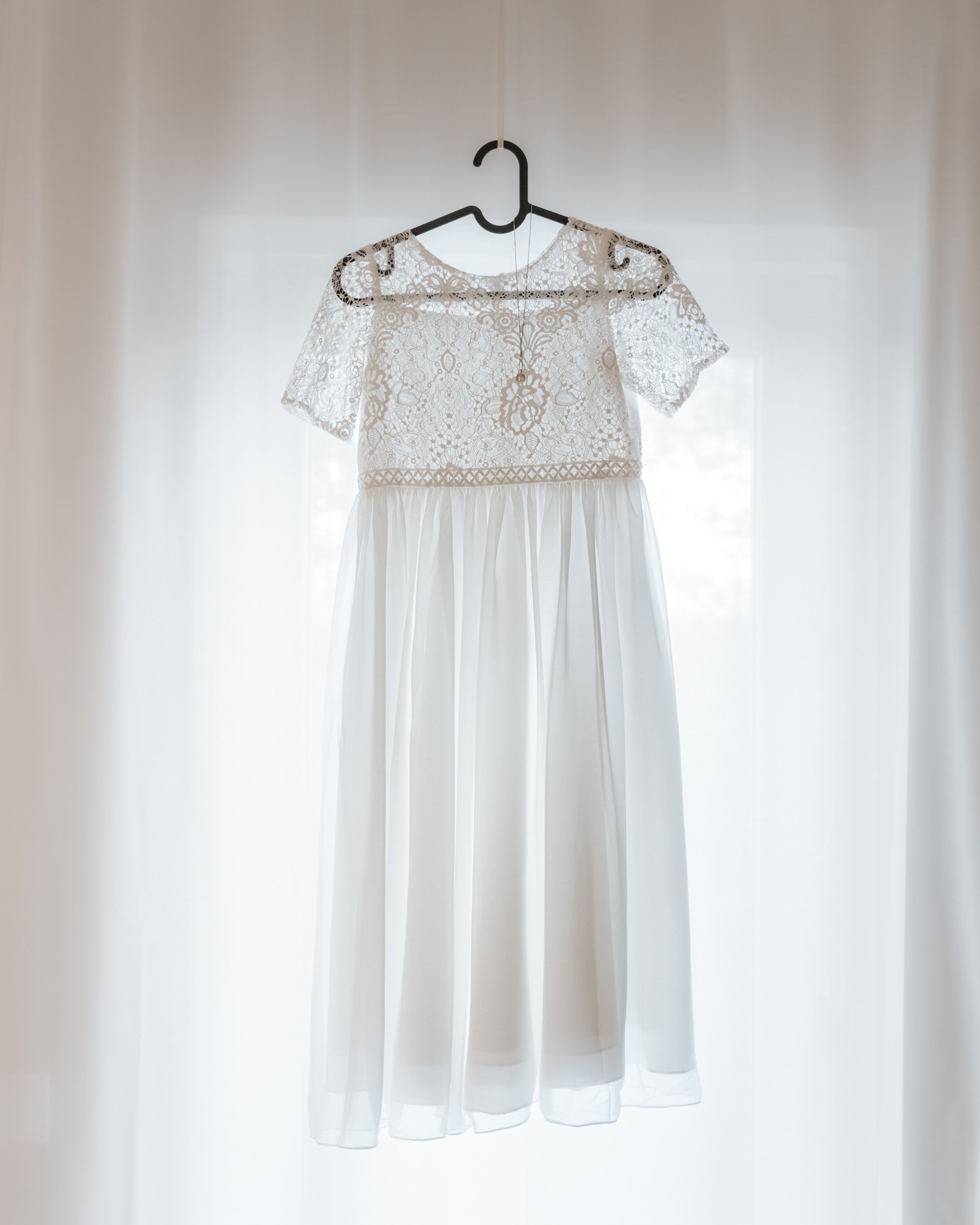 A white dress for a girl with lace bodice and short lace sleeves hanging on a black hanger against a light background.
