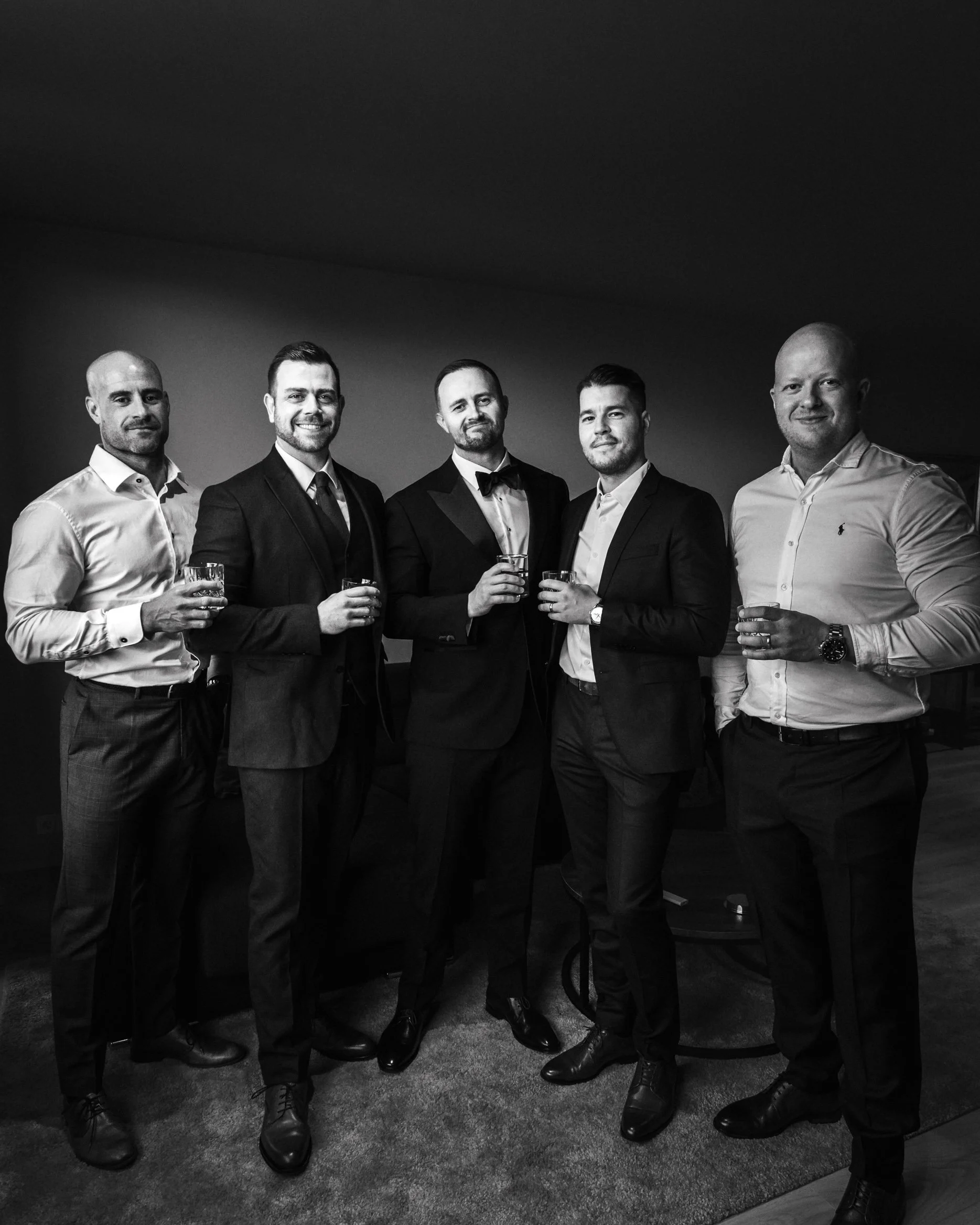 Five men in formal attire holding drinks, standing together indoors.