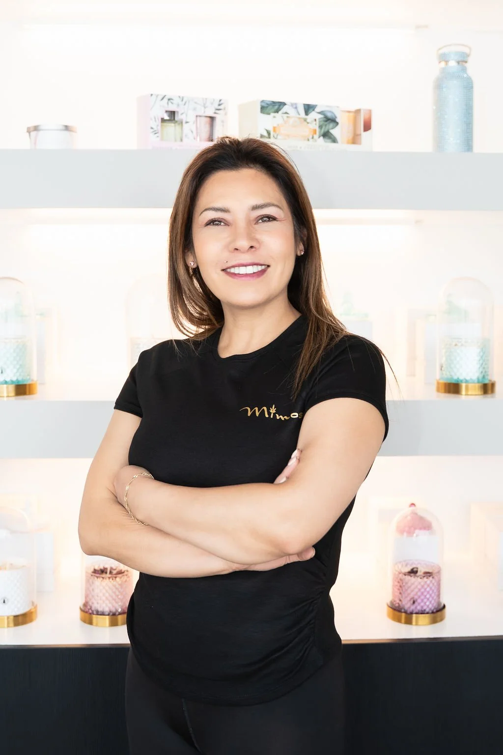 A woman with brown hair and a black T-shirt standing confidently with arms crossed in front of a display of colorful decorative containers.