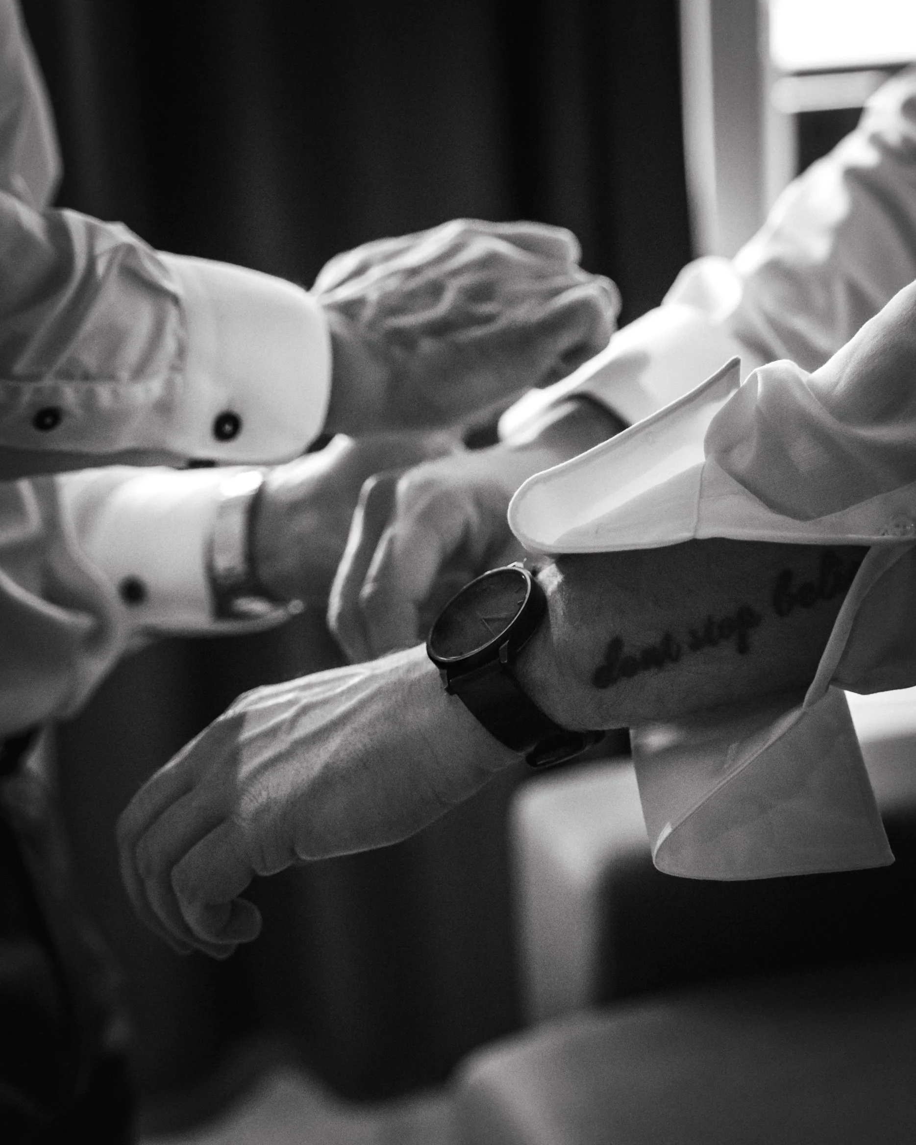 Hands of two individuals exchanging a small item, both wearing long-sleeve shirts, one with a sleeve rolled up and a watch on the wrist, in a black and white photo.
