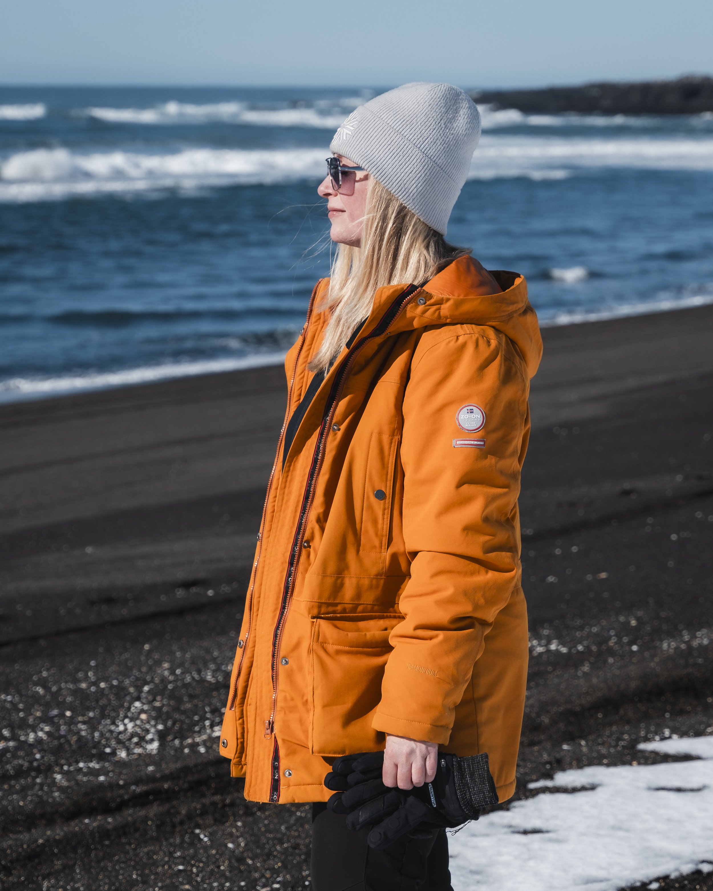 A woman standing on a black sand beach wearing an orange winter jacket, gray knit beanie, sunglasses, and black gloves, with the ocean and waves in the background.