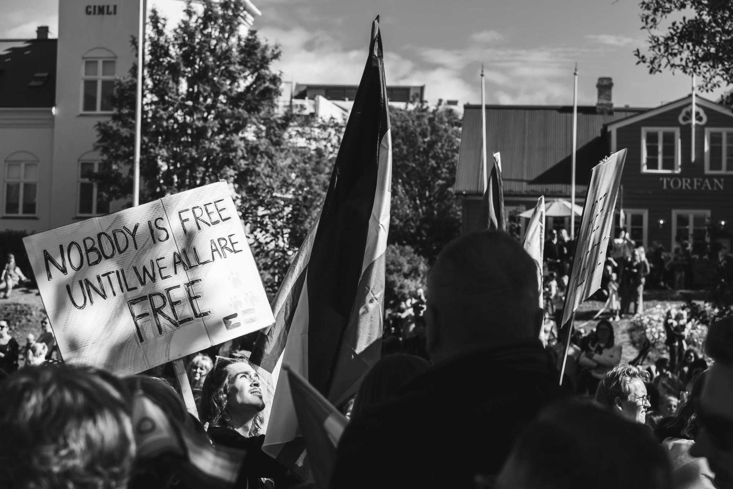 A crowd of people at a protest or rally, with some holding signs and flags. One sign reads 'Nobody is free until we all are free.' There are buildings and trees in the background, and it appears to be daytime with partly cloudy skies.