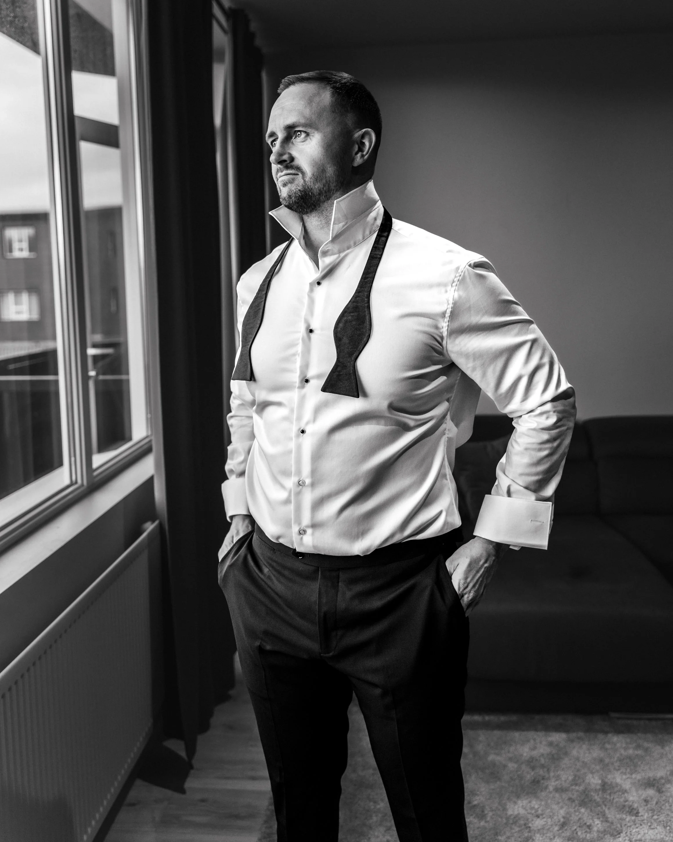 A man in formal attire, with a white shirt and dark pants, standing near a window in a modern living room, looking out contemplatively.