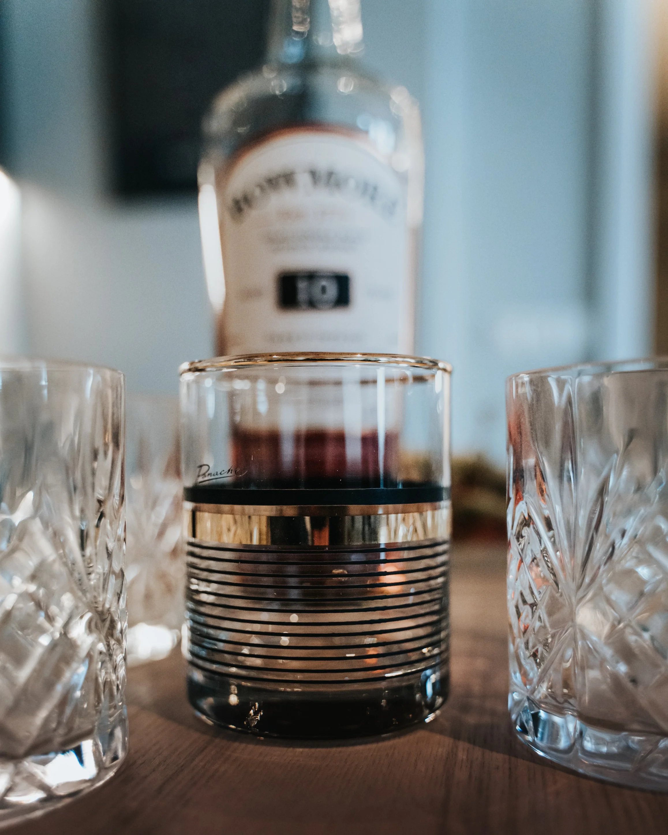 A bottle of Proper No. Twelve Irish whiskey in the background with a glass of dark liquor in the foreground, flanked by two cut crystal glasses on a wooden surface.