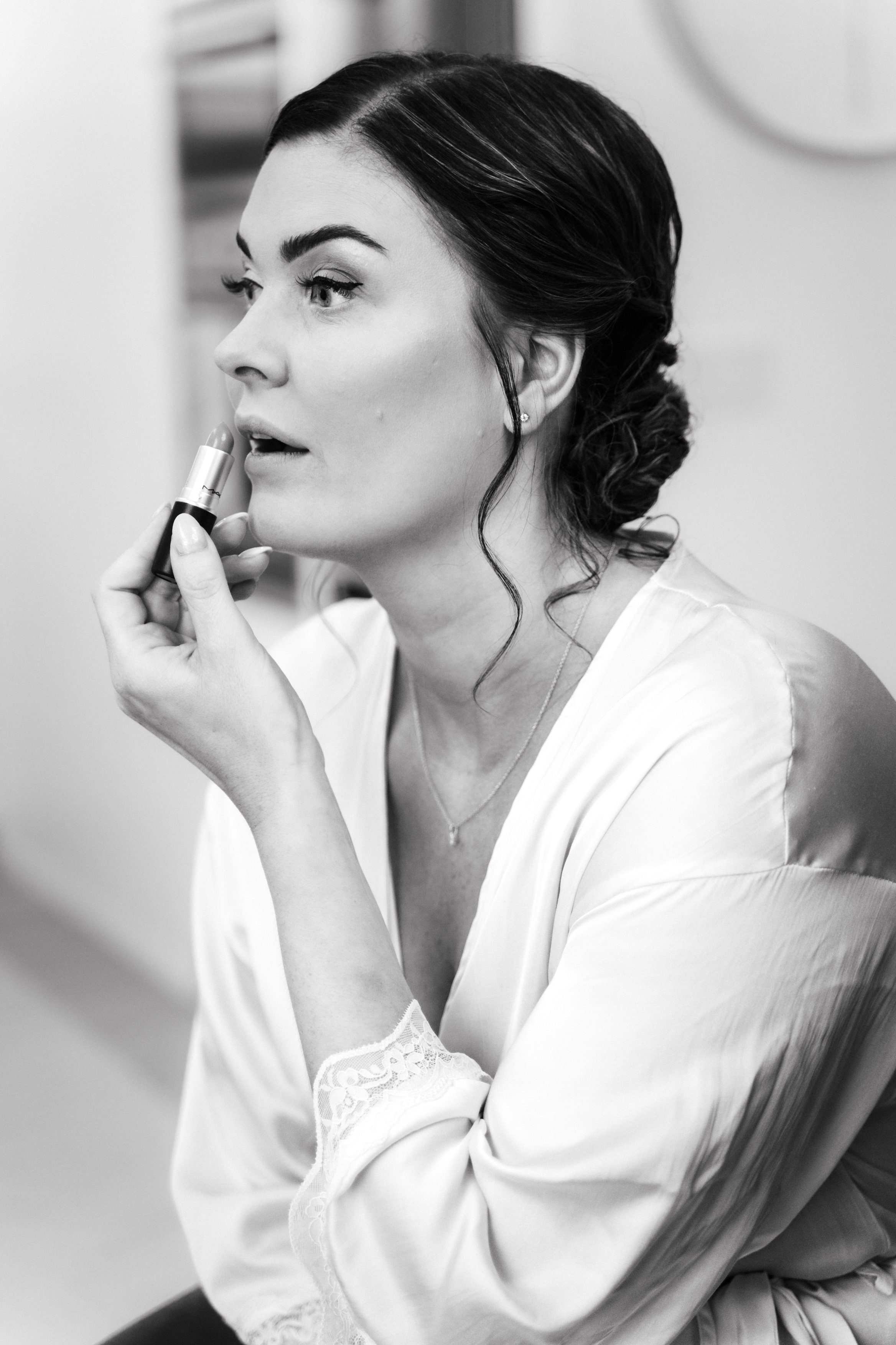 Woman applying lipstick in front of mirror, wearing a satin robe and jewelry, with her hair styled in an updo.