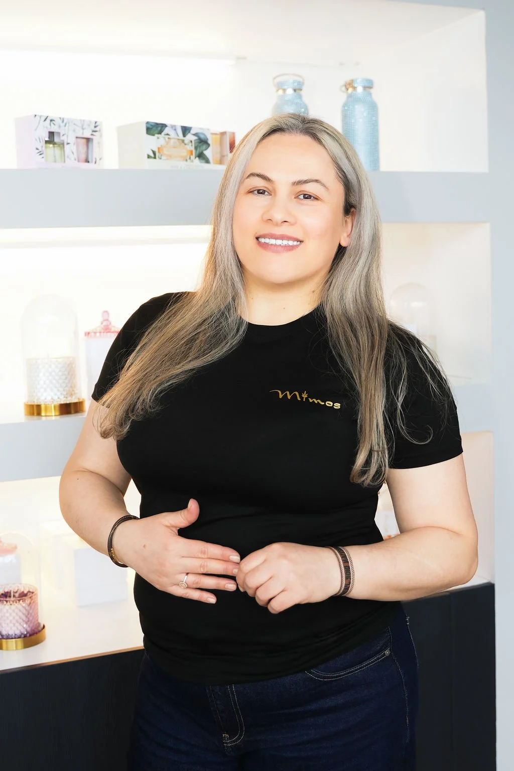 A woman with long gray hair smiling at the camera stands indoors against a backdrop of shelves decorated with candles and vases.