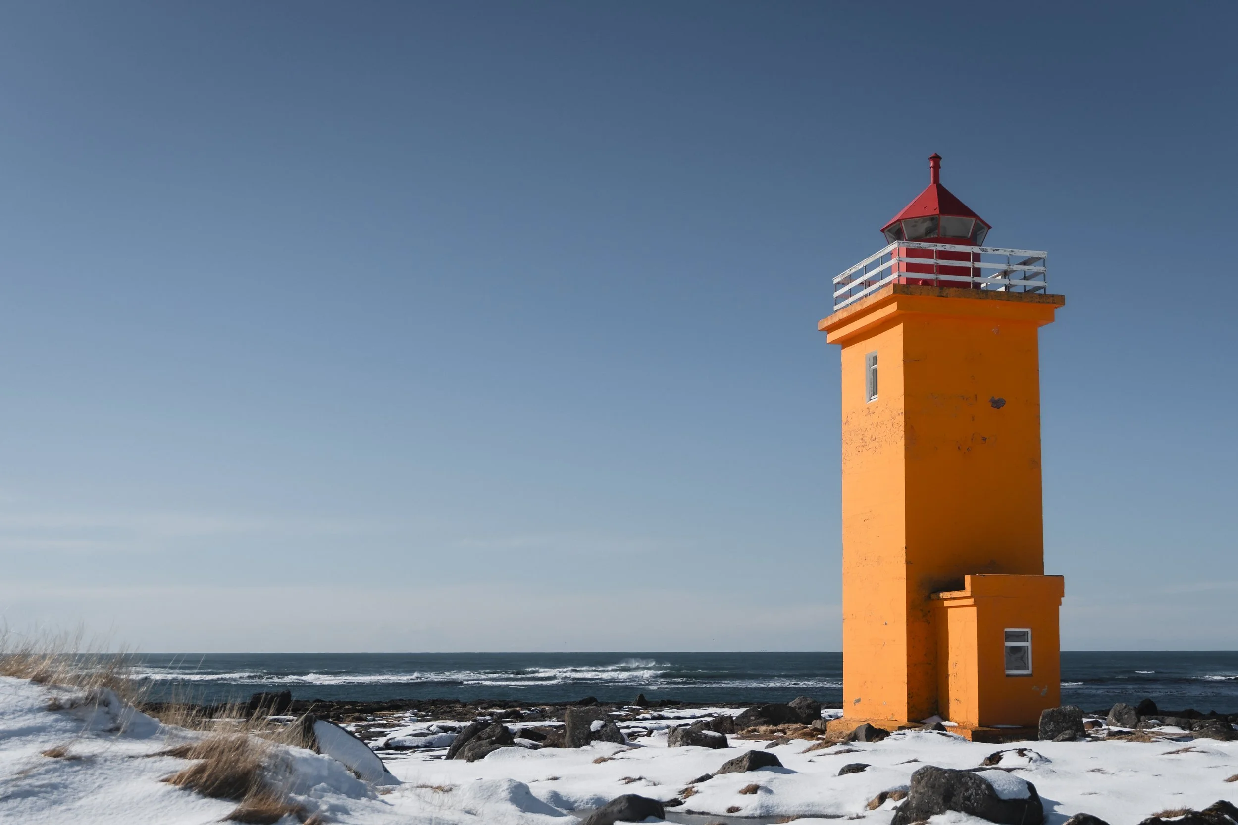 Orange lighthouse on rocky, snow-covered shoreline with the ocean in the background and a clear blue sky.