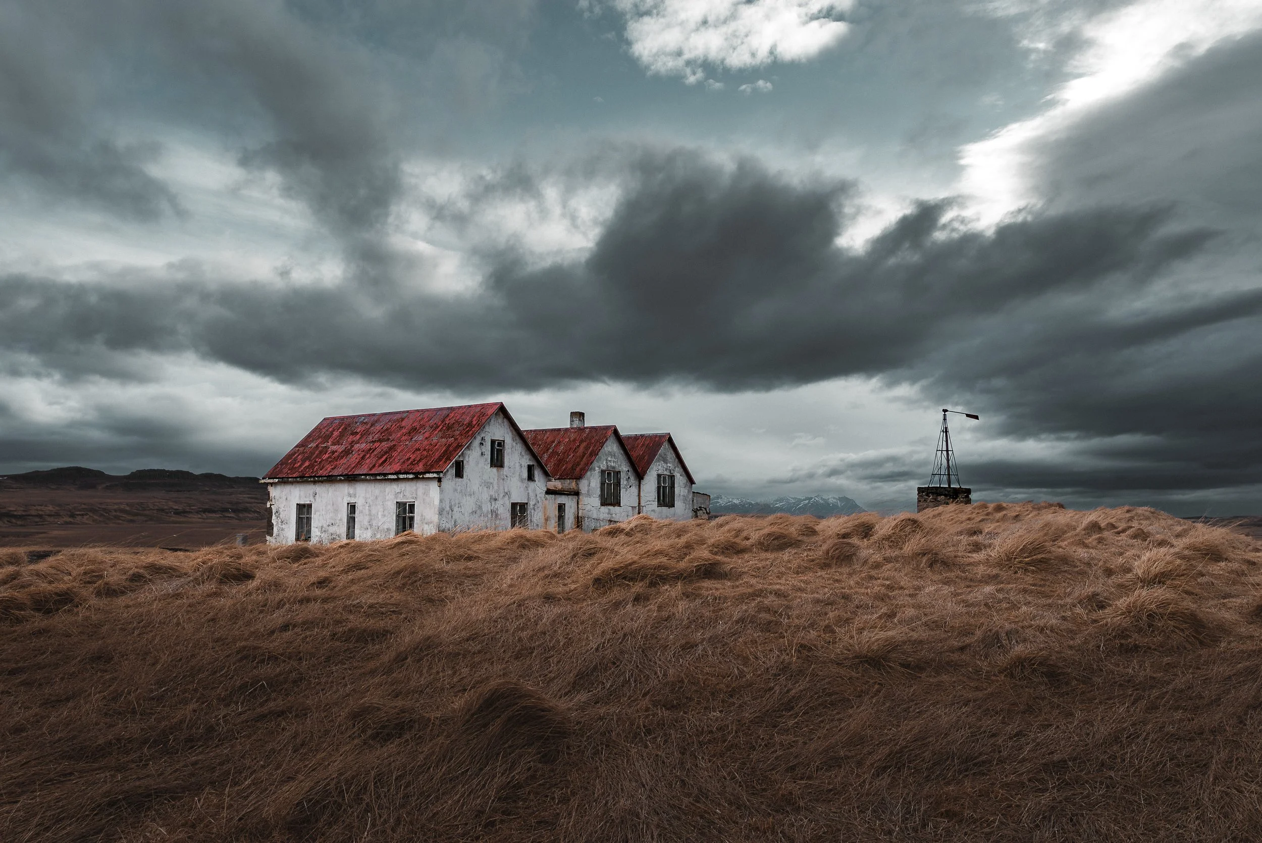 A weathered white house in rural Iceland, with a rusted red roof stands in a field of dry, brown grass. Dark storm clouds fill the sky, and mountains are visible in the background, with a small windmill to the right of the house.
