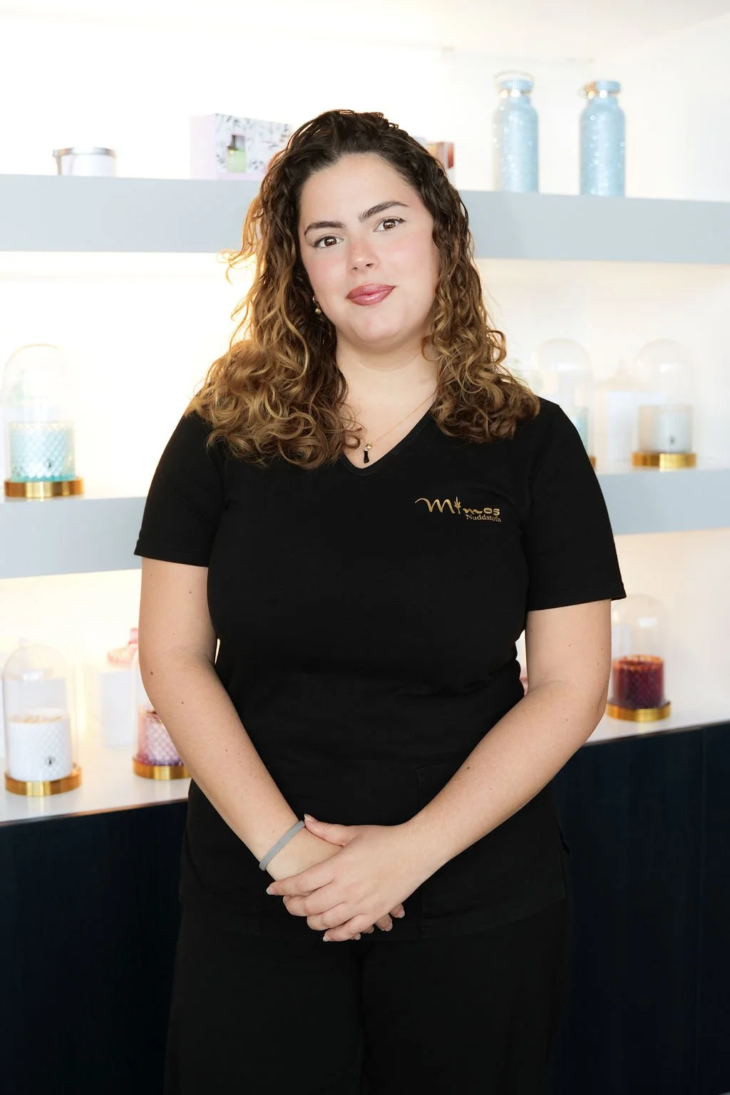 A woman with curly brown hair wearing a black t-shirt with gold writing, standing in front of a display of glass containers filled with colored objects.