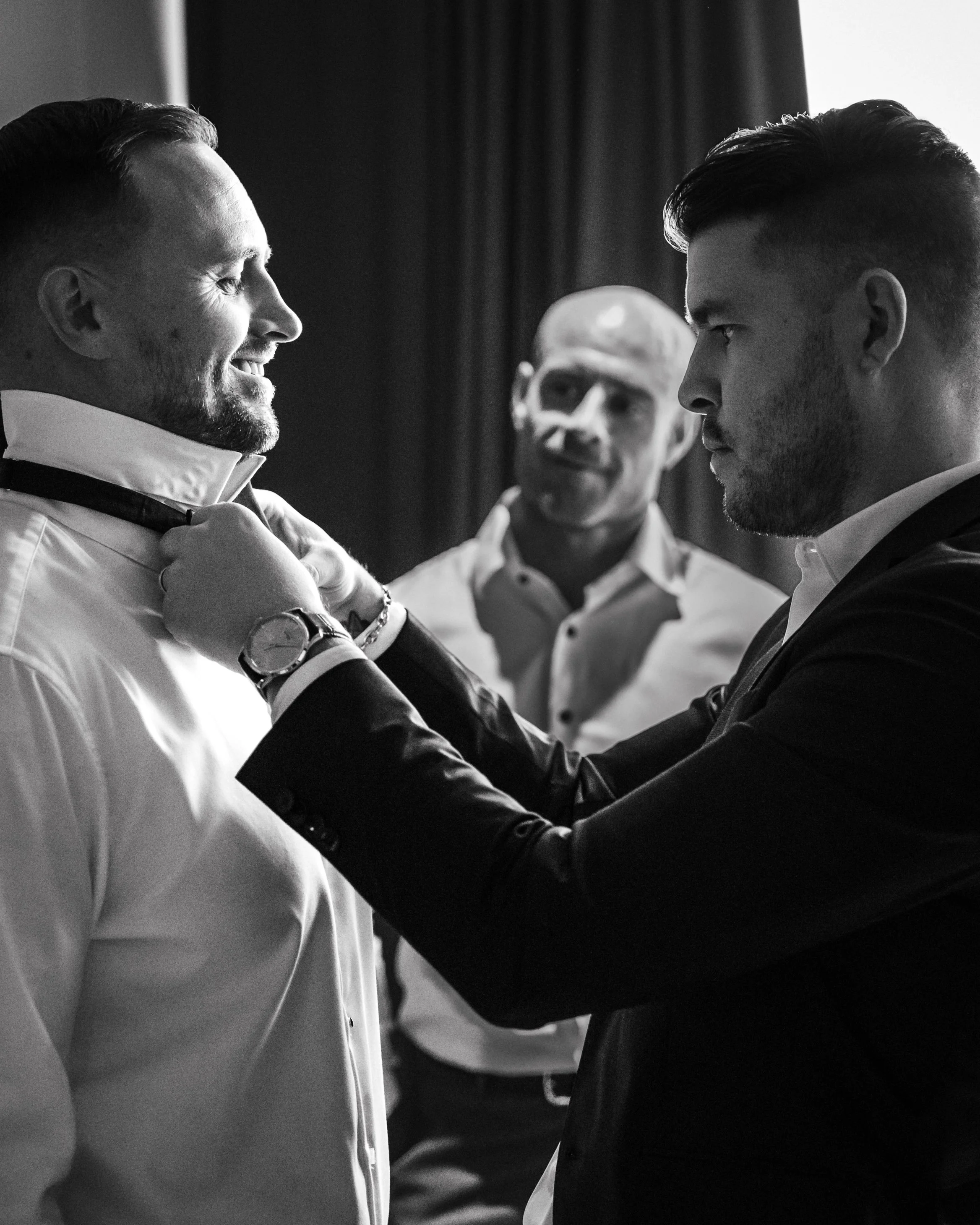 Three men in formal attire, one helping another adjust his tie, in a black-and-white photo.