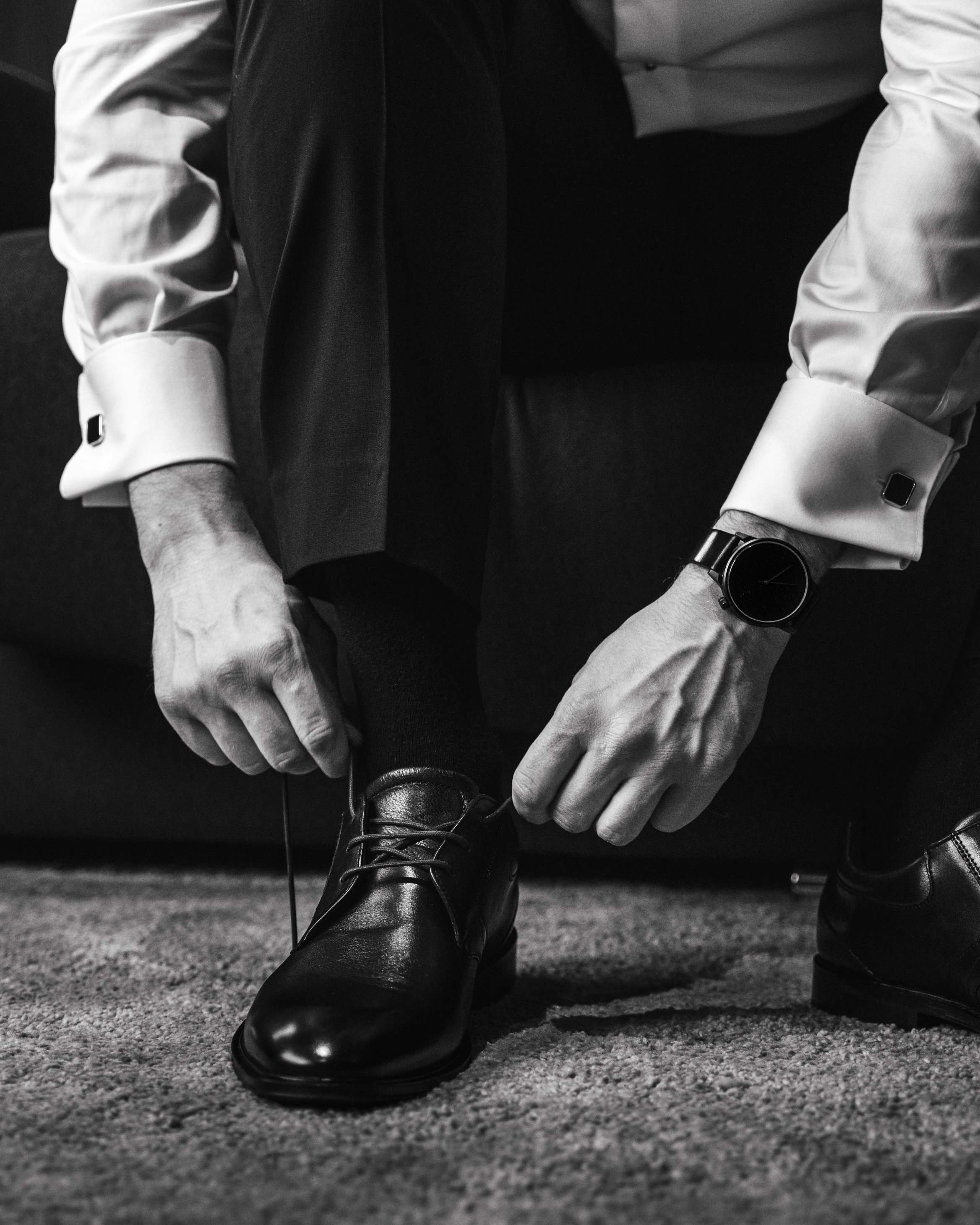 A man dressed in formal attire is sitting on a couch, tying his black leather shoe. He is wearing a white shirt with cufflinks, a suit, and a wristwatch.