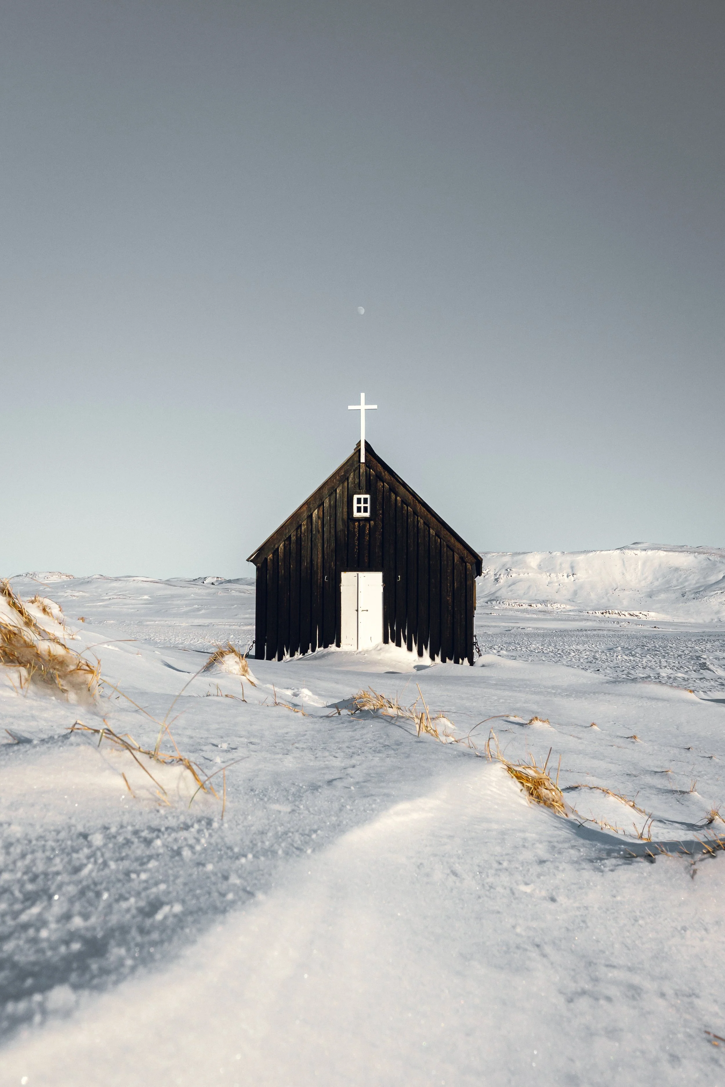 A small black church stands alone in a vast, snow-covered landscape, framed by soft light and endless sky. Above it, the faint presence of the moon lingers—quiet, distant, and still. There is a sense of calm here that feels almost sacred, where silen