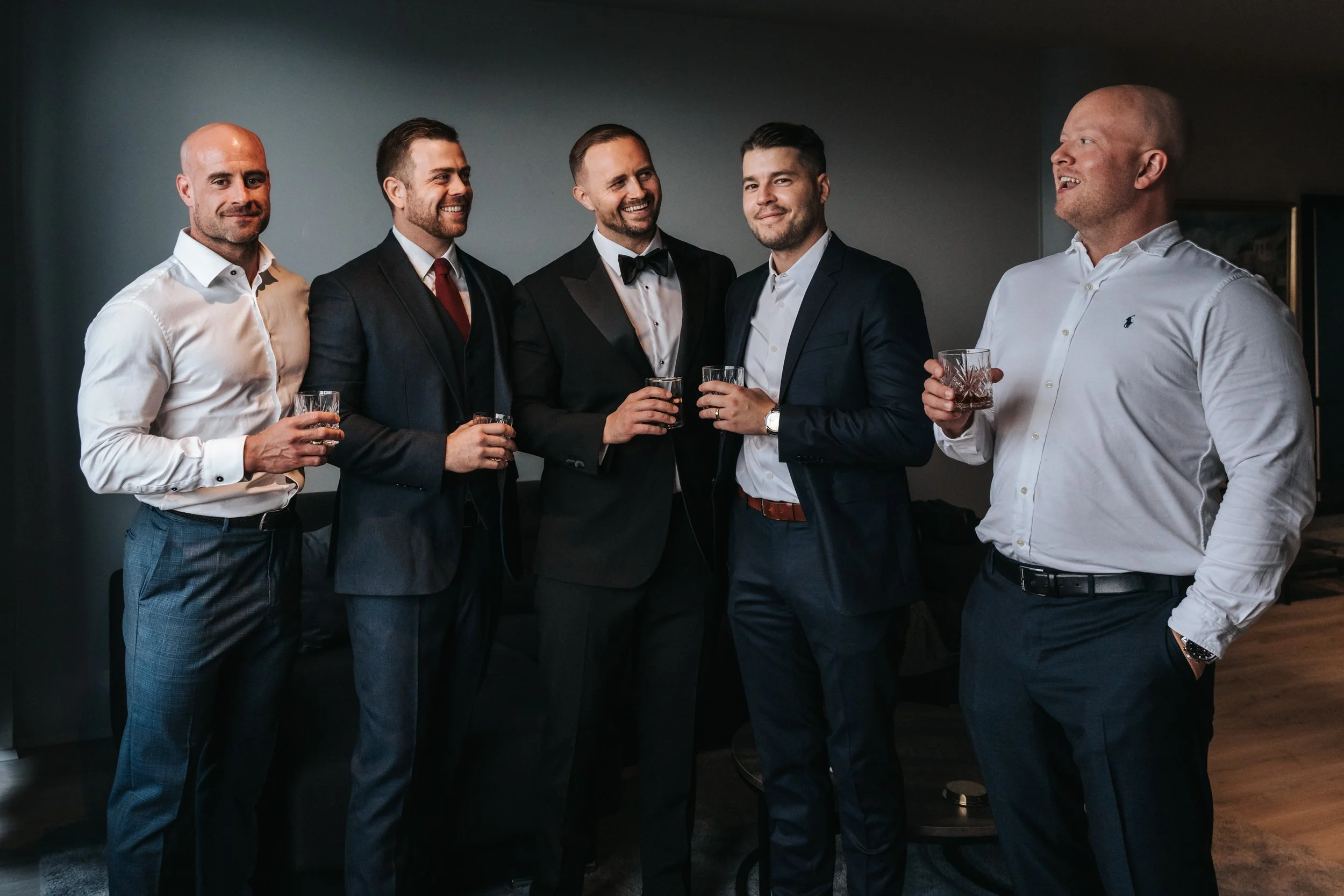 Group of six men in formal attire with drinks in hand, standing indoors, engaging in conversation, smiling, at a social gathering or celebration.