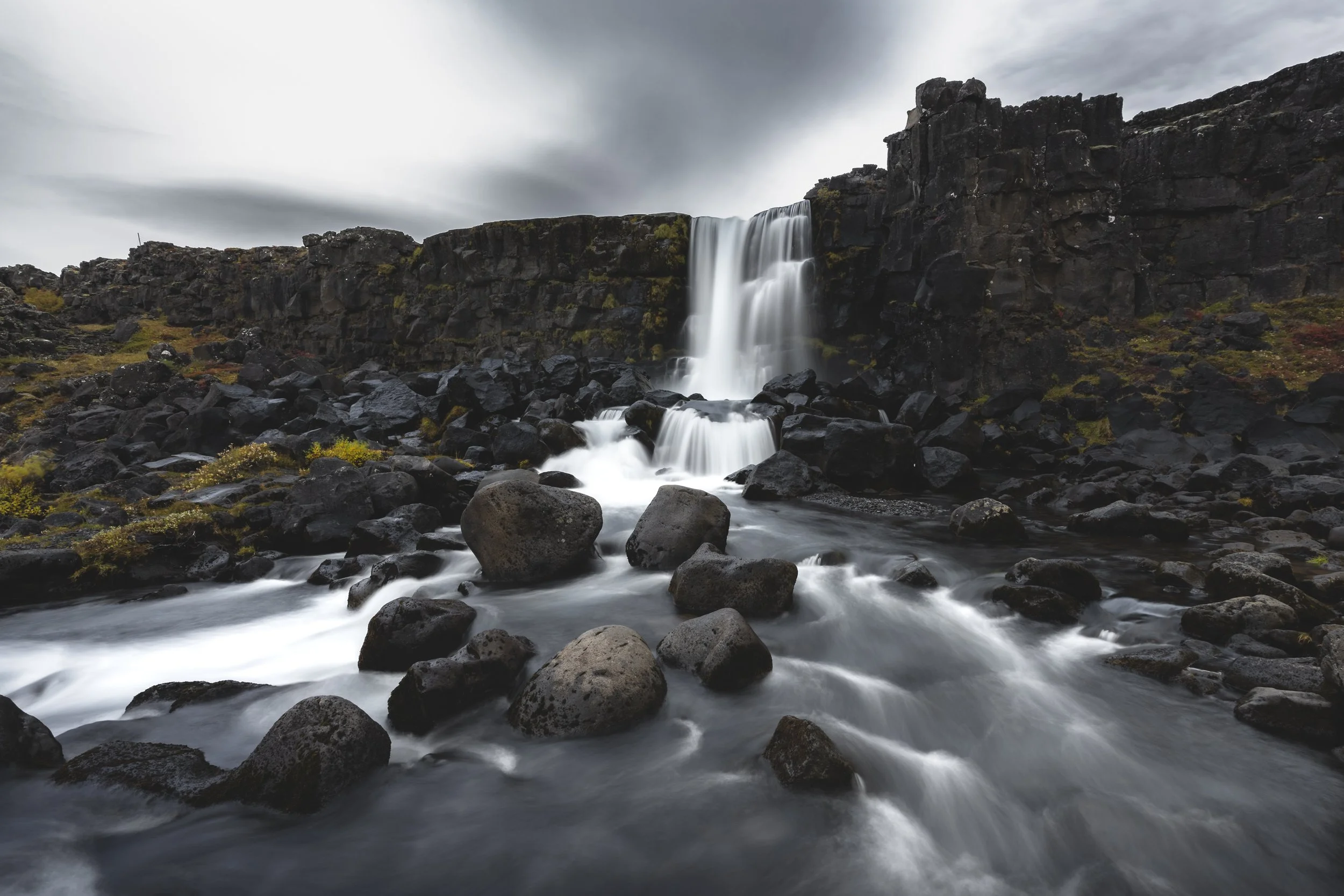A waterfall flowing over black volcanic rocks surrounded by a rocky riverbed under a cloudy sky.