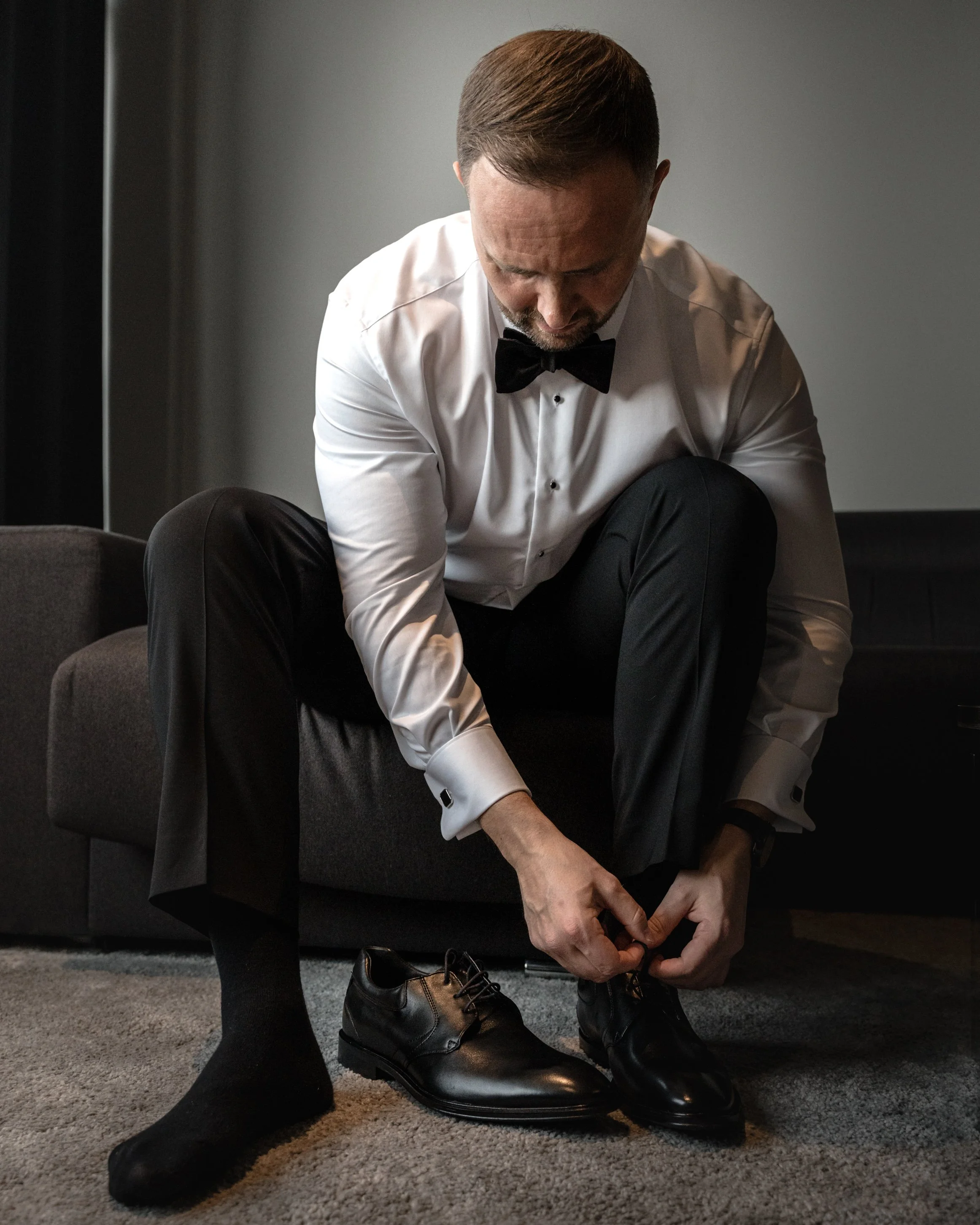 A man in a tuxedo with a bowtie sitting on a couch, tying his black dress shoes on a gray carpet.