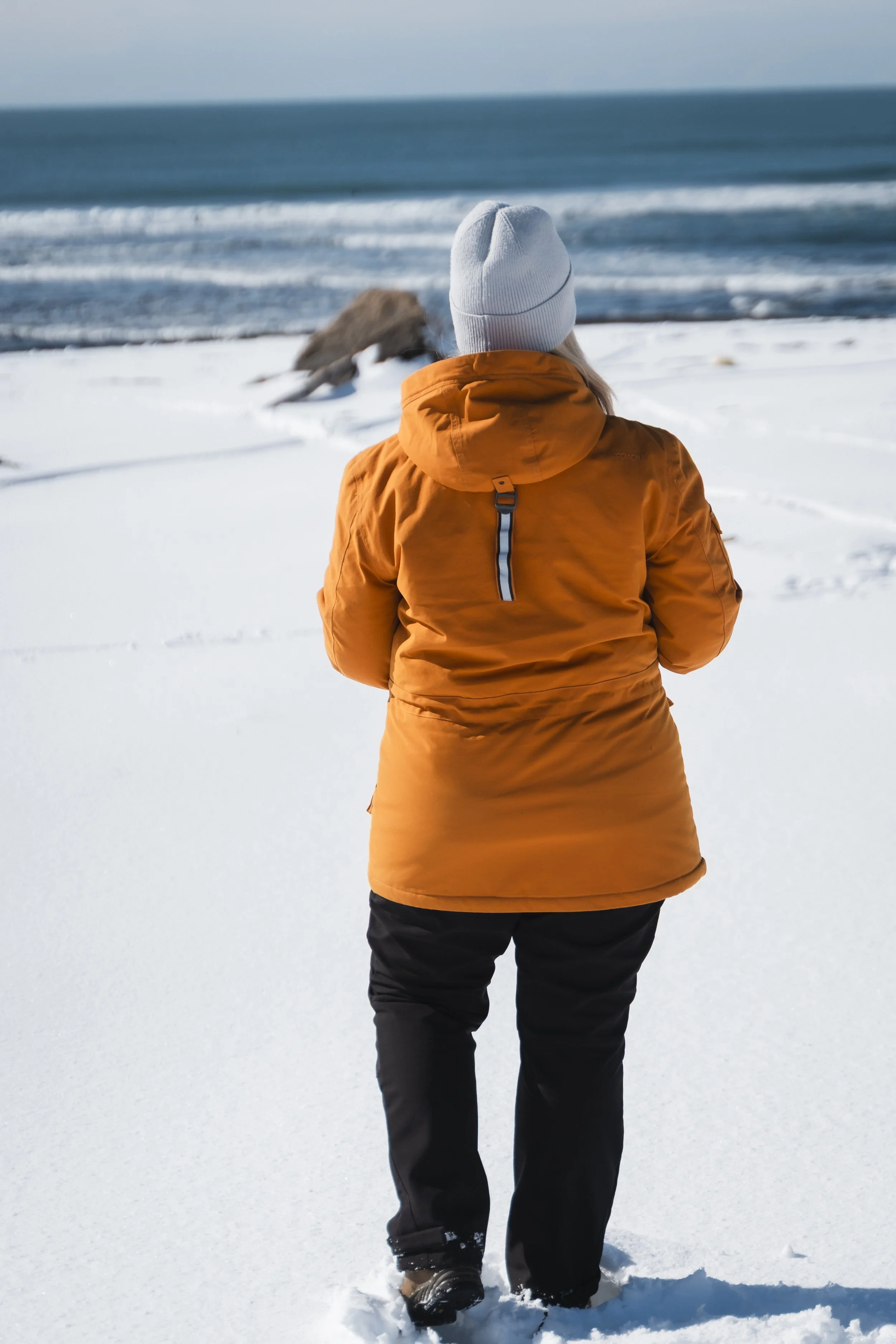 Person in an orange winter coat and gray beanie standing on snow-covered ground, facing the ocean with waves, rocks, and a cloudy sky in the background.