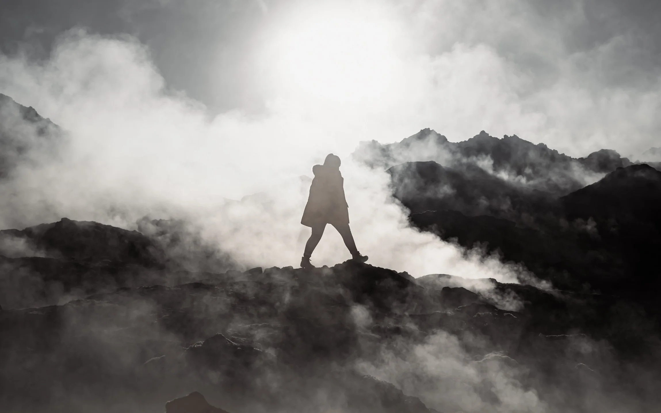 A person walking on rocky terrain surrounded by clouds and mist with mountain peaks in the background.
