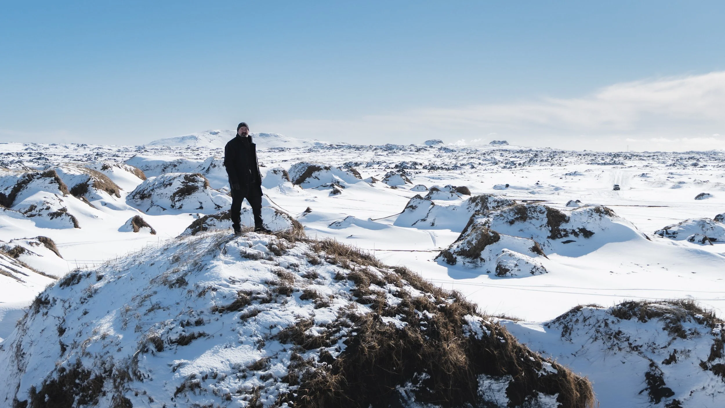 A man dressed in dark clothing standing on a snow-covered hill with a vast snowy landscape and blue sky in the background.