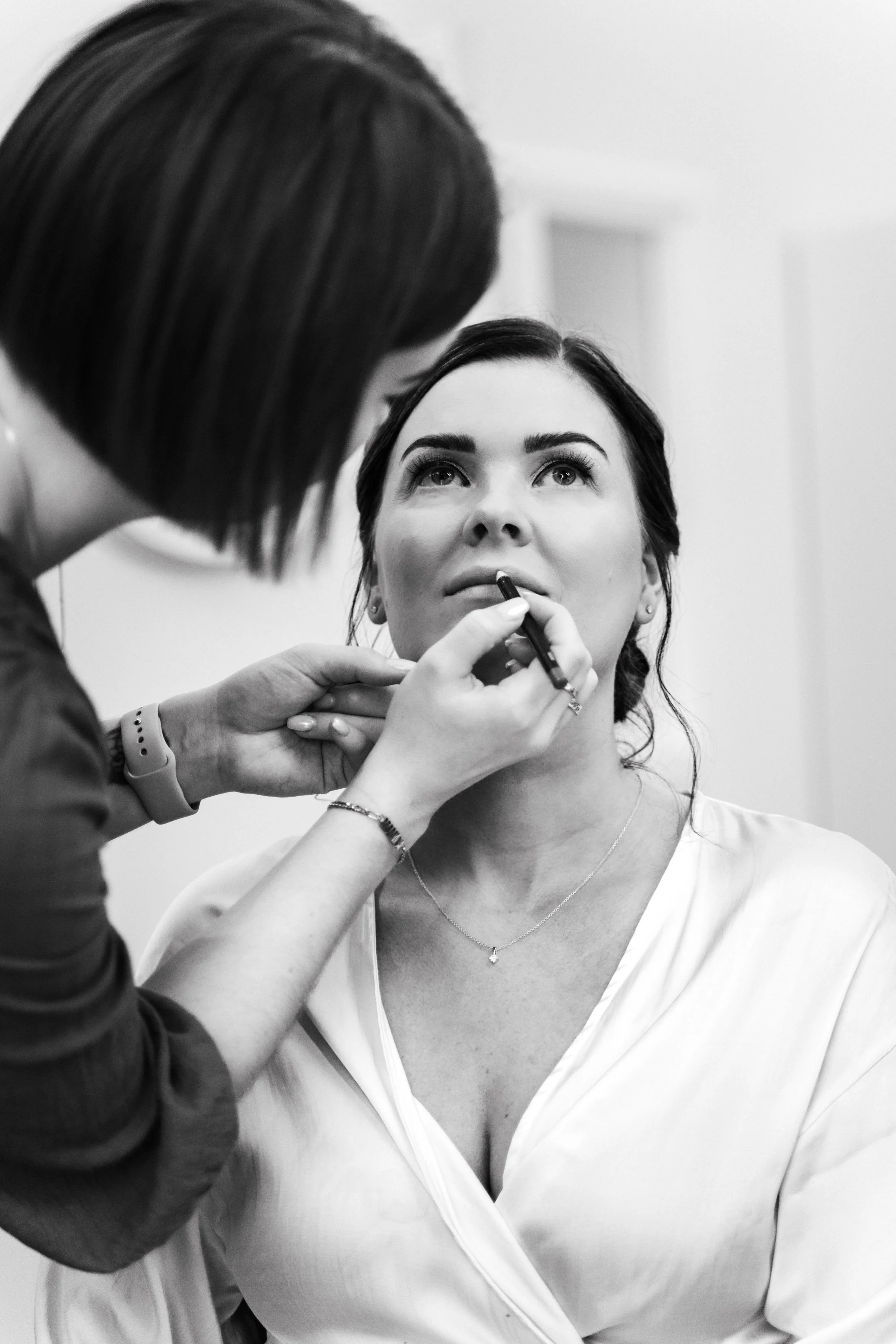 A woman with dark hair and makeup being assisted by a makeup artist applying makeup to her lips.
