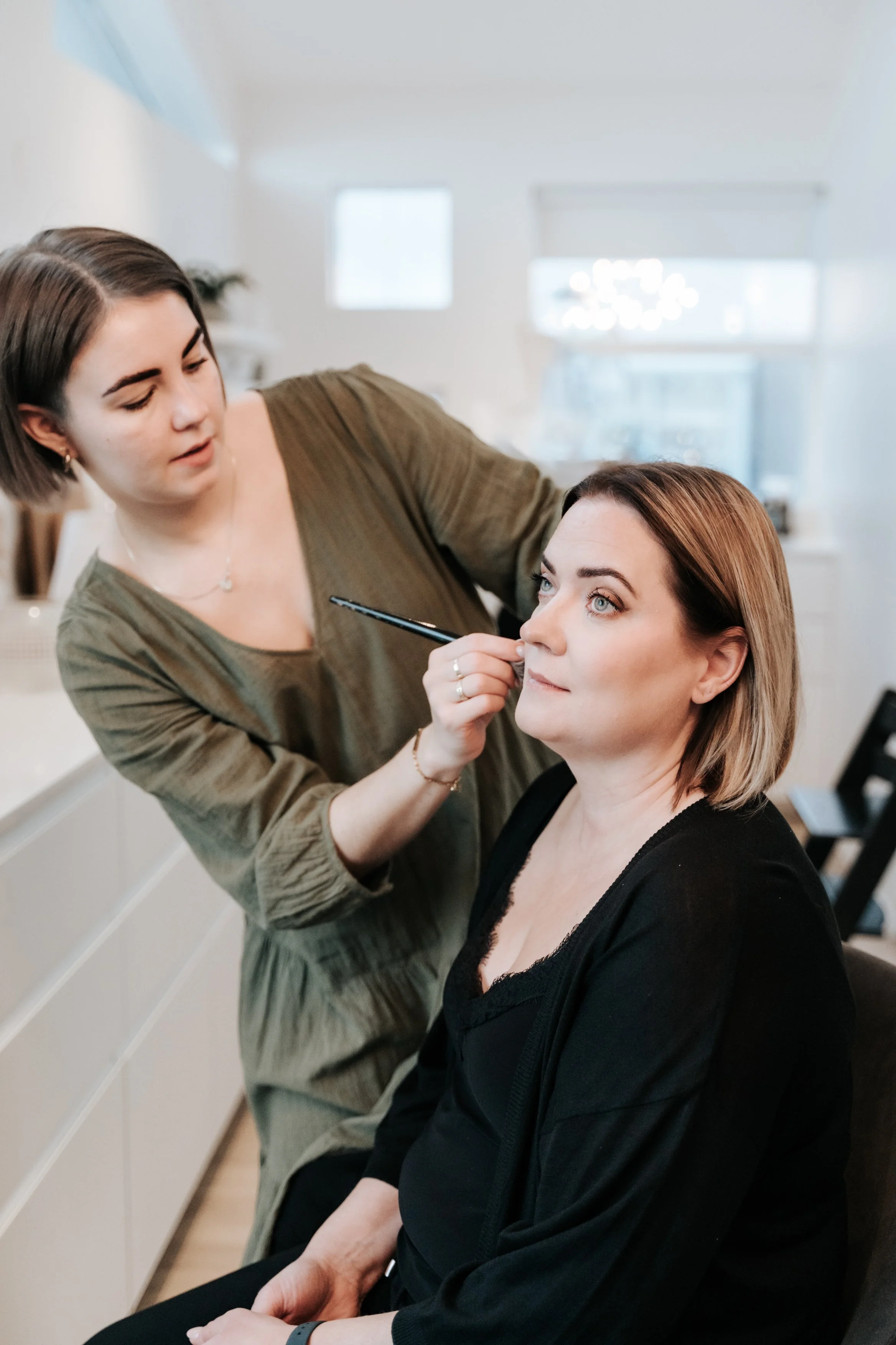 A woman sitting in a makeup chair getting her makeup done by a makeup artist in a bright, modern room.