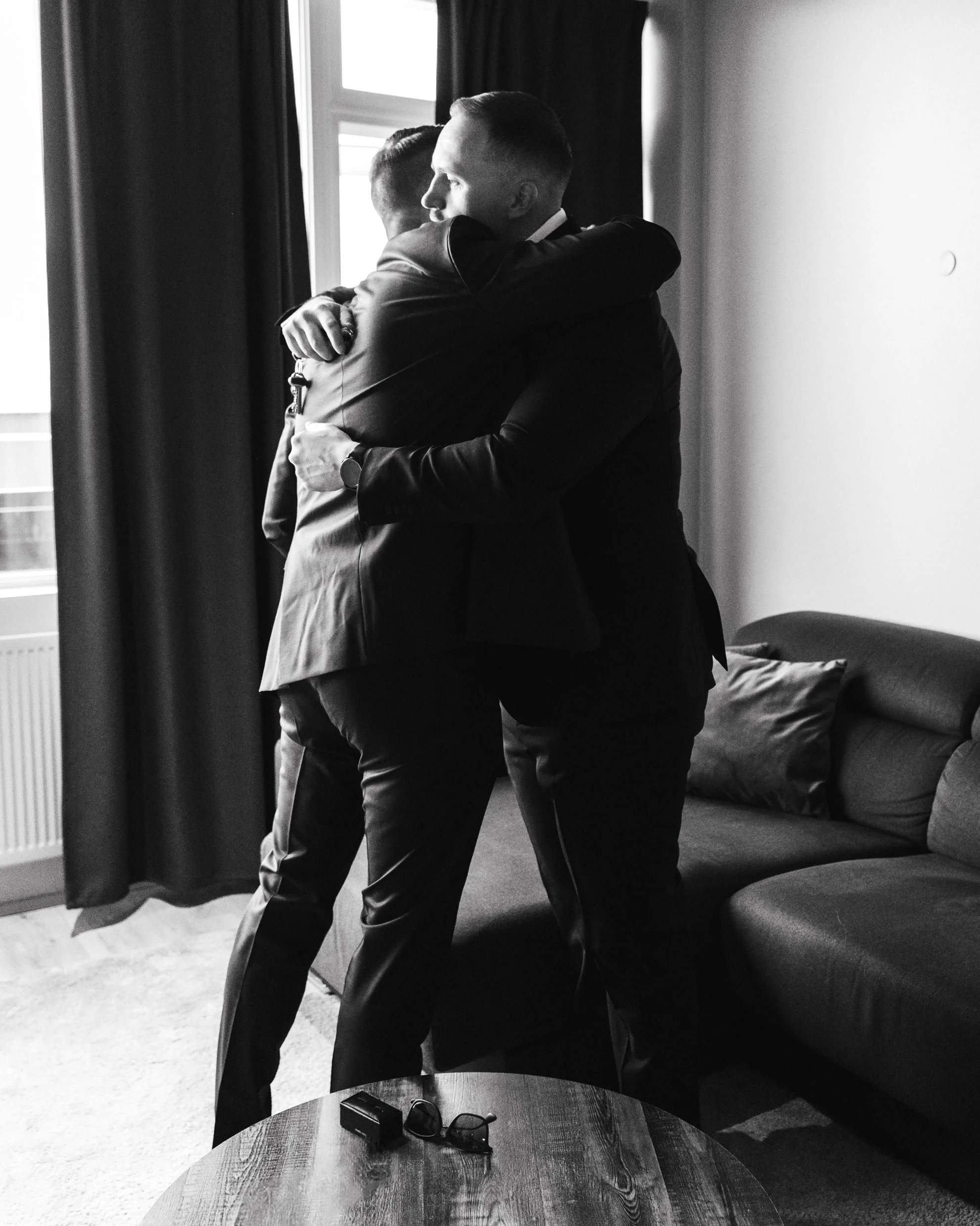 Two men in suits hugging in a room with a window, sofa, and a wooden table with glasses and a small device on it.
