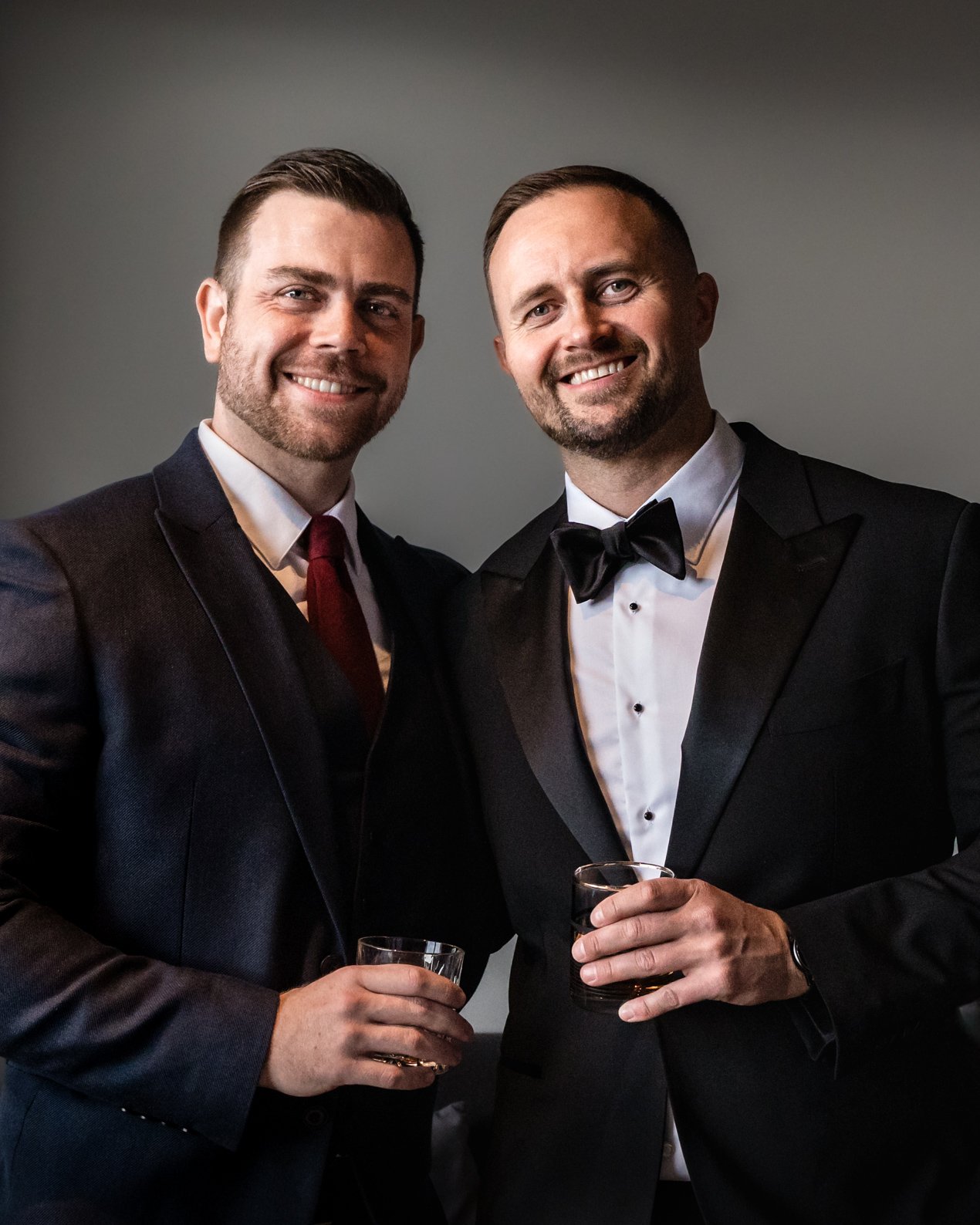 Two men in tuxedos holding glasses of whiskey at a formal event.