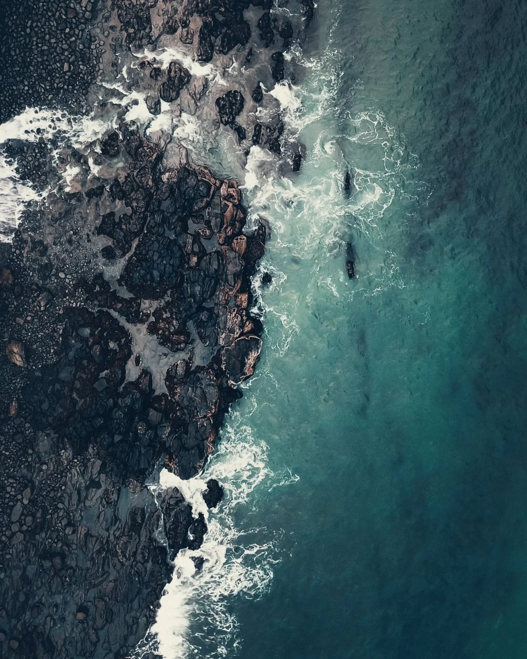 An aerial view of a rocky shoreline with waves crashing against dark rocks and greenish-blue ocean water.
