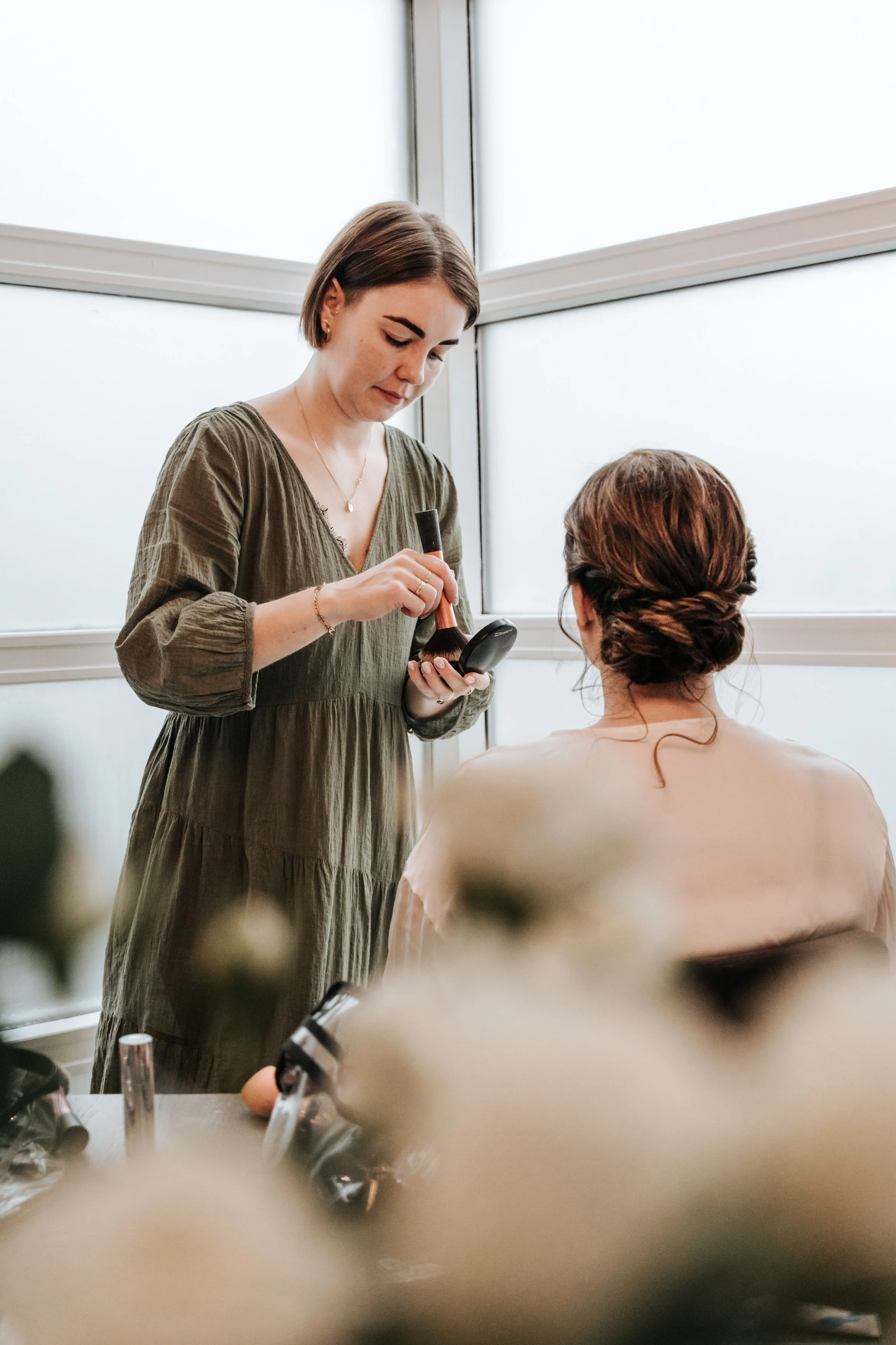A woman in an olive green dress applies makeup to another woman with her back to the camera, in front of large frosted windows indoors.