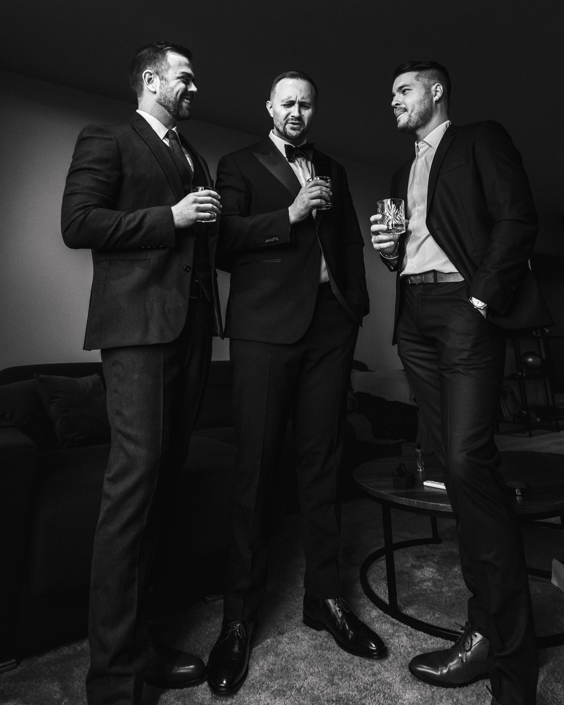 Three men in suits holding drinks, standing indoors, engaged in conversation, with a sofa and table in the background. Black and white photo.