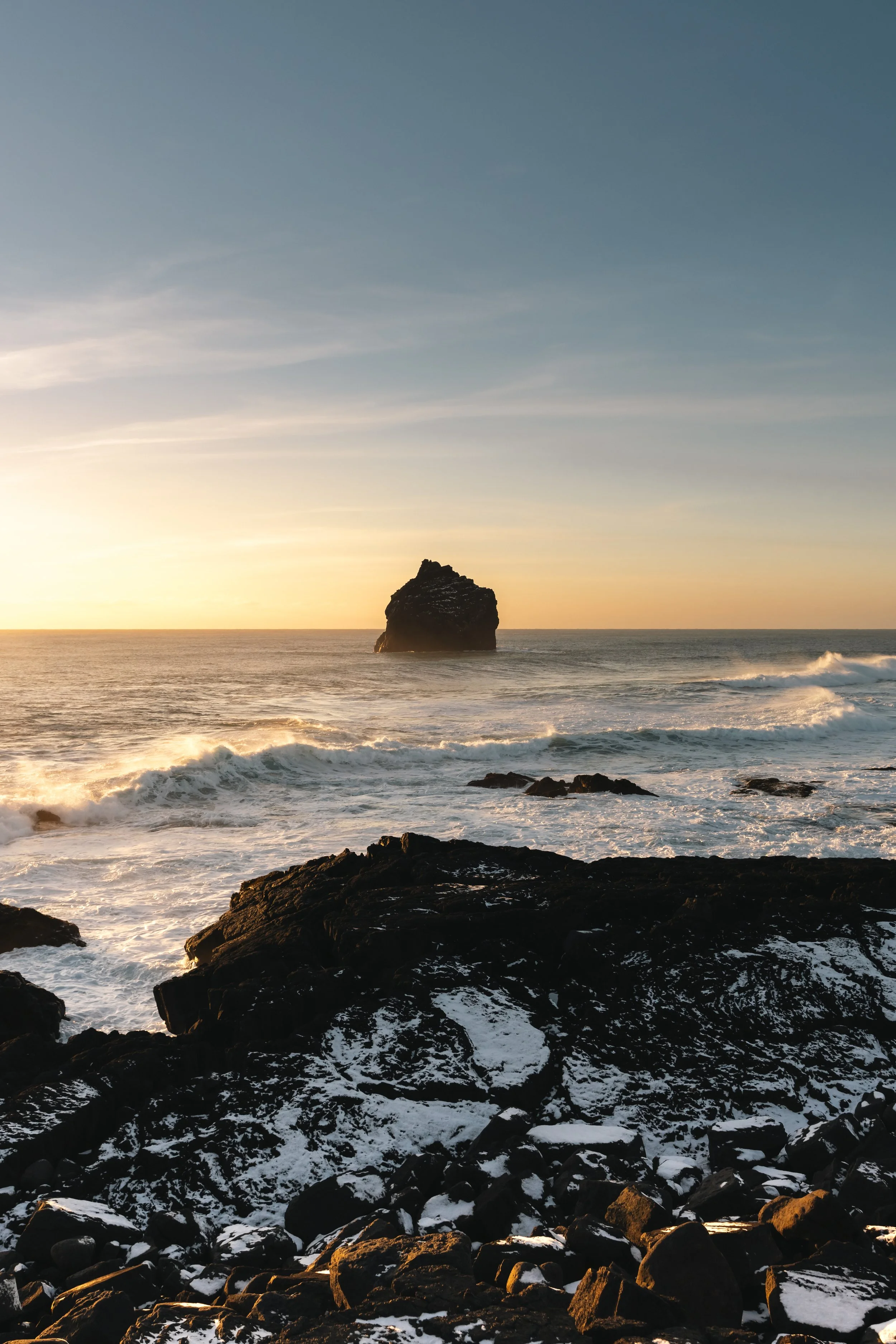 Seaside view at sunset with waves crashing onto rocky shore and a large rock formation in the distance.