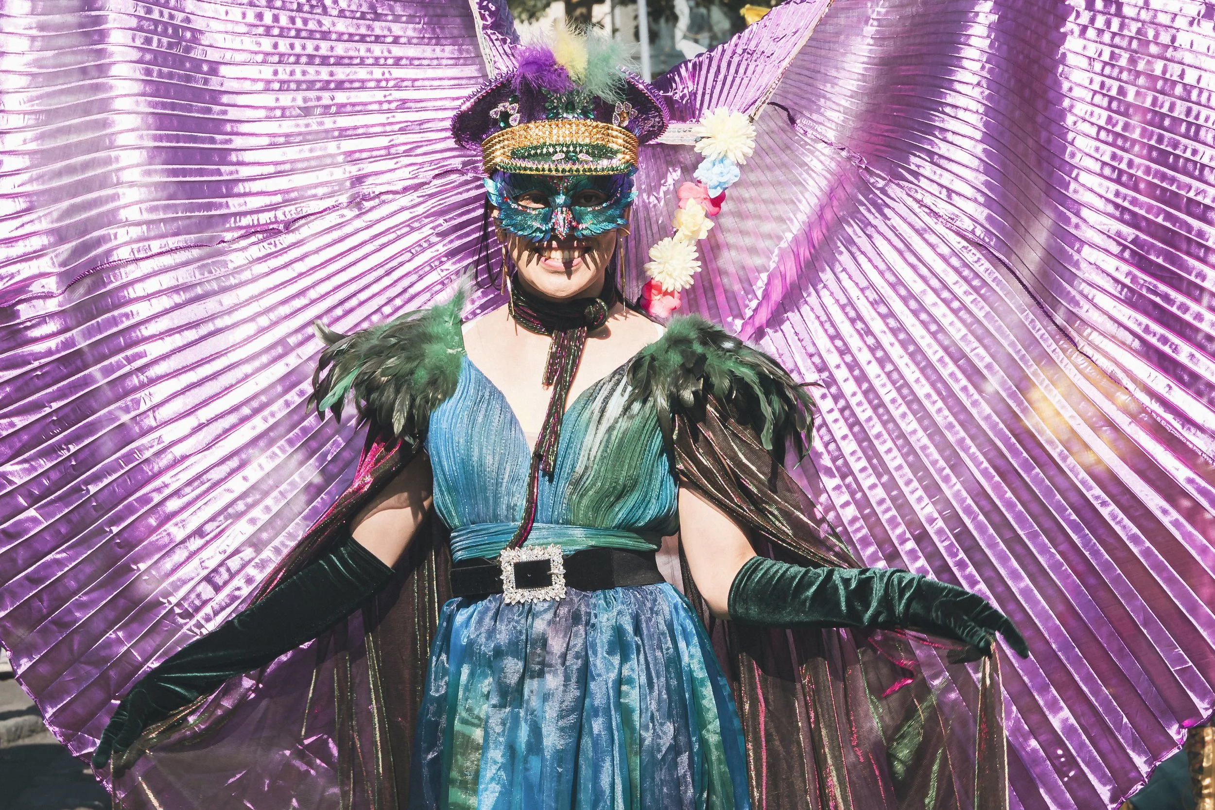 Person in colorful, feathered costume with a mask, standing in front of large pink and purple metallic fan backdrop.