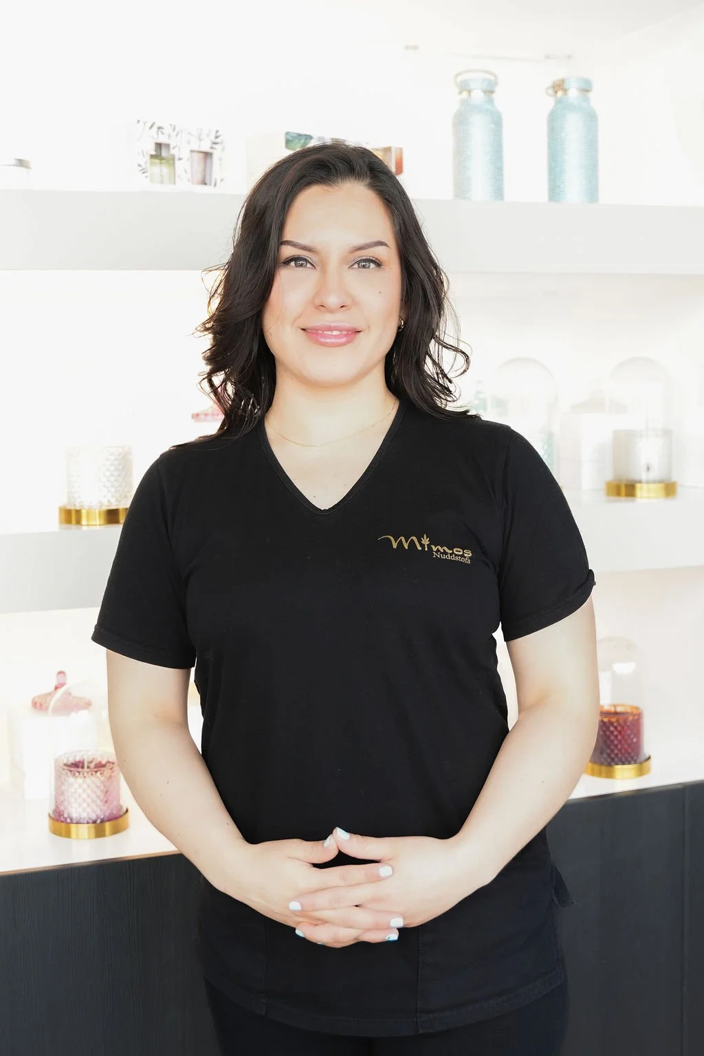 A woman with dark wavy hair, wearing a black T-shirt, standing in a modern room with decorative items on white shelves behind her.