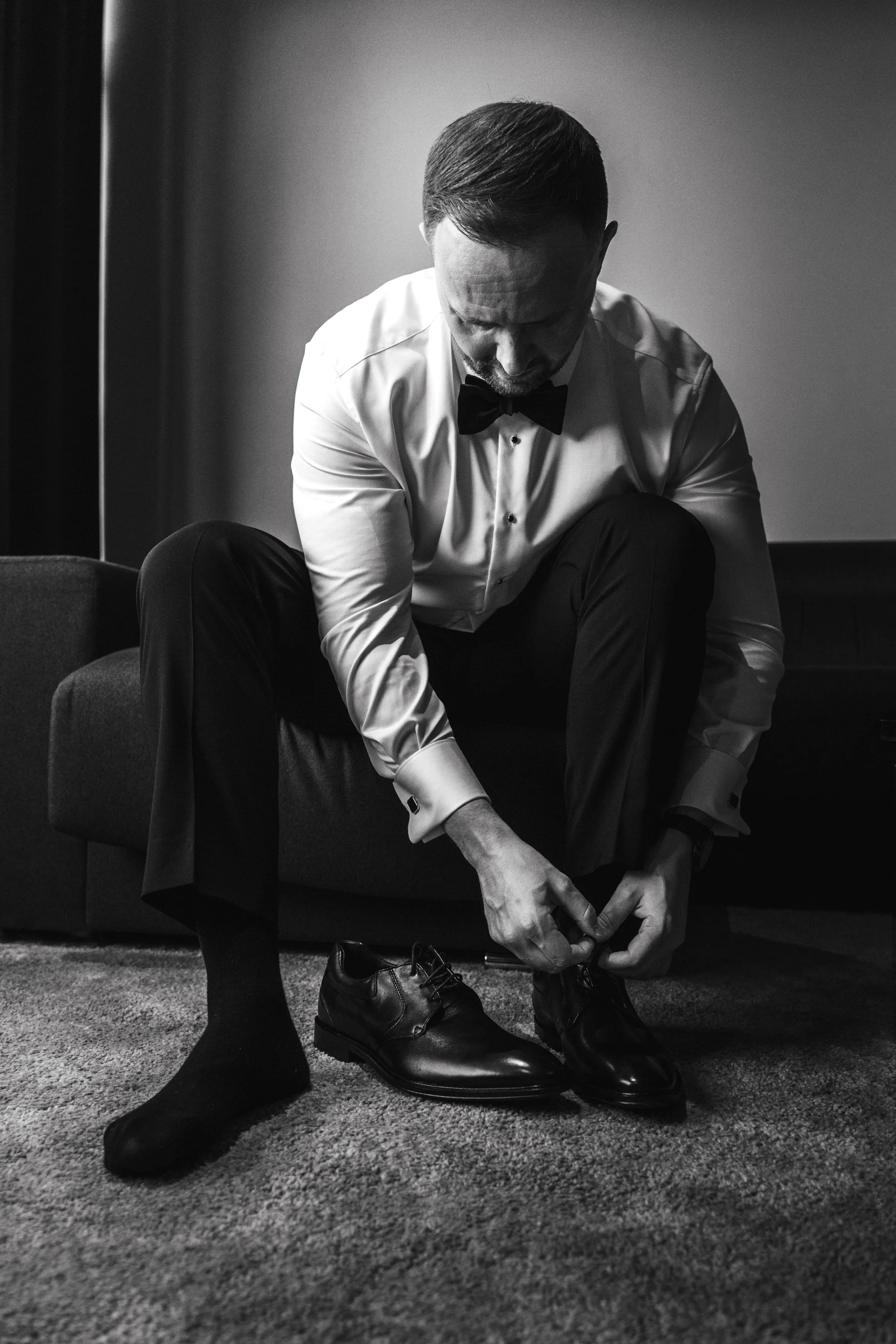 A man in a tuxedo and socks is sitting on a sofa, tying his dress shoes in a dimly lit room.