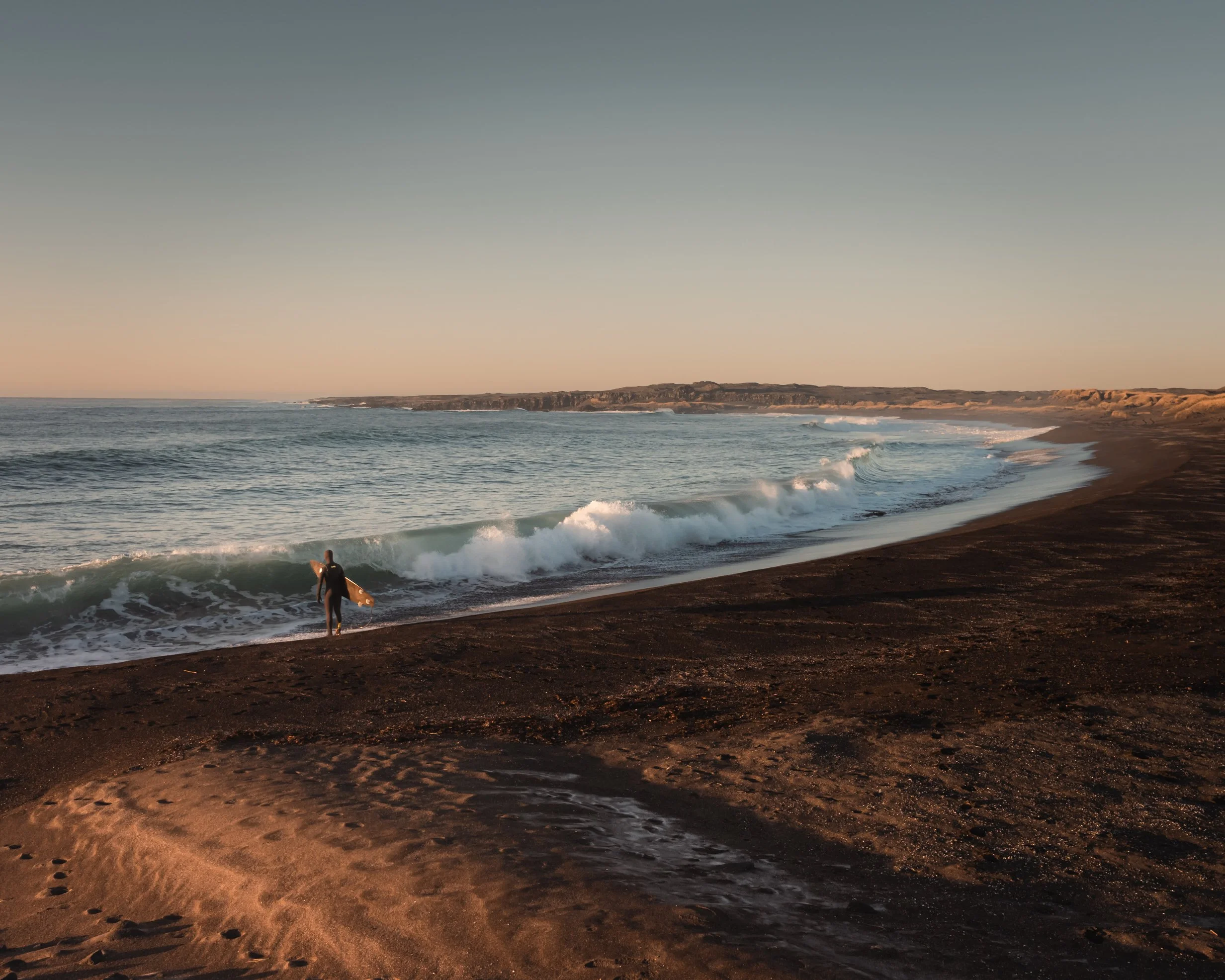 A person with a surfboard walking into the ocean on a sandy beach during sunset.