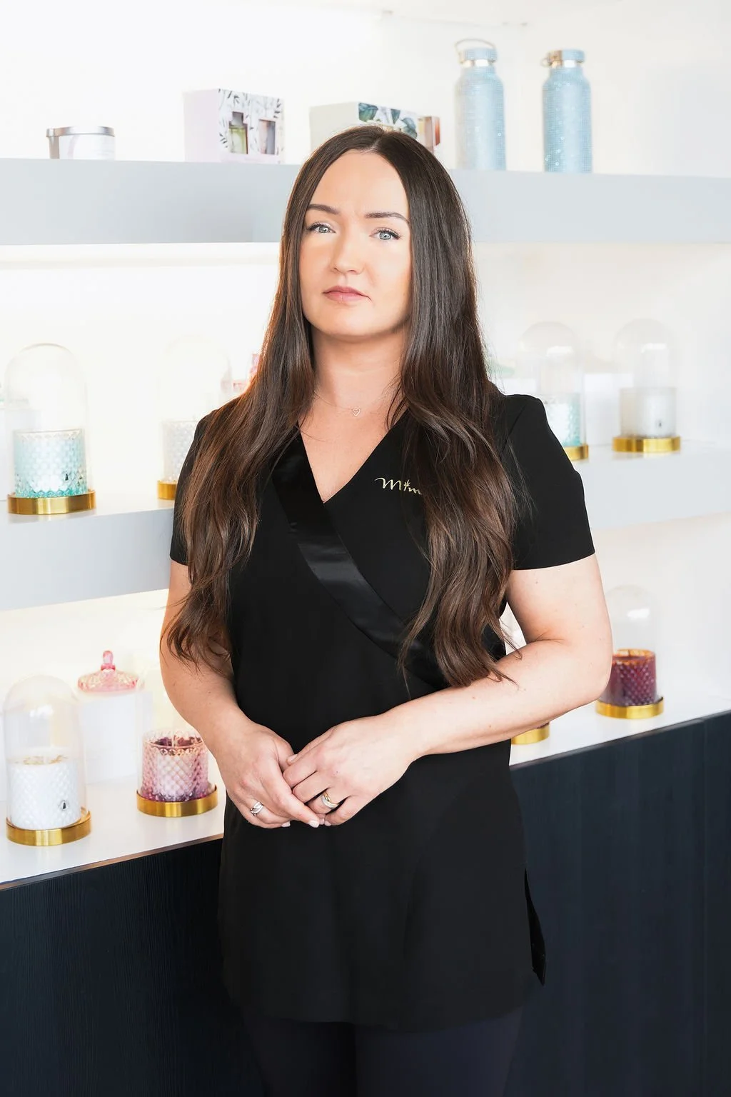 A woman with long brown hair and fair skin wearing a black uniform standing in front of a display shelf with decorative candles and bottles.