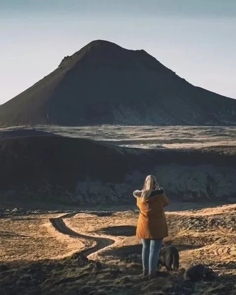 A person with a dog standing on a dirt path in front of a mountain at sunset or sunrise.