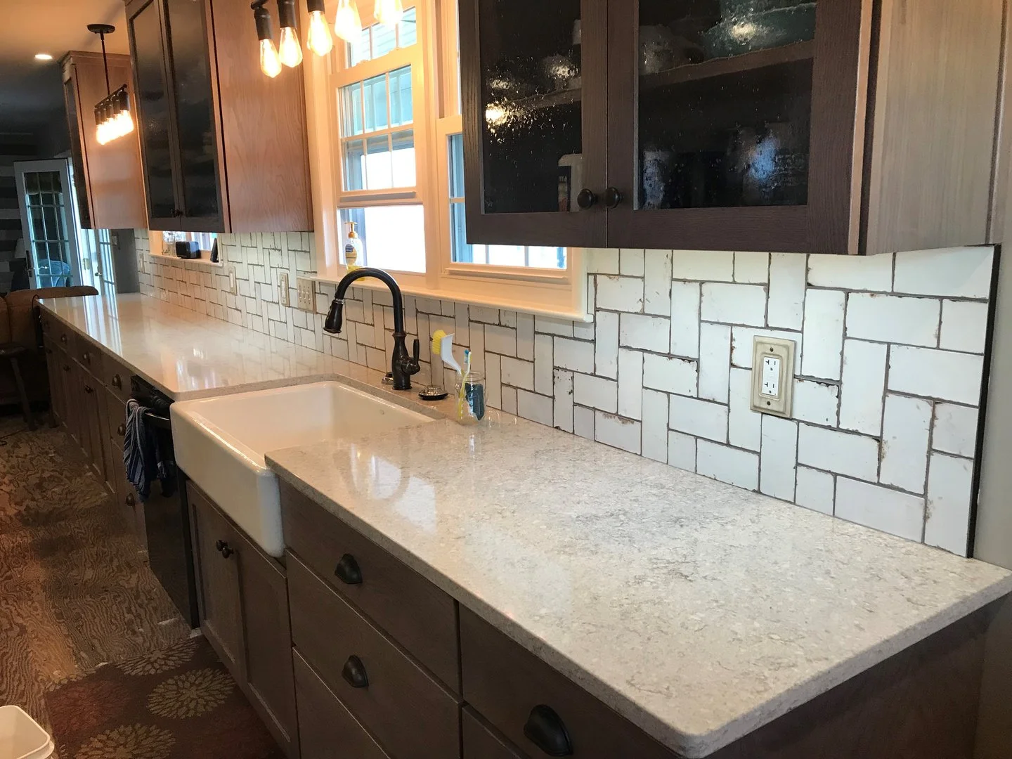 Modern kitchen with white countertops, farmhouse sink, dark fixtures, dark wood cabinets, and white tile backsplash under warm pendant lighting.