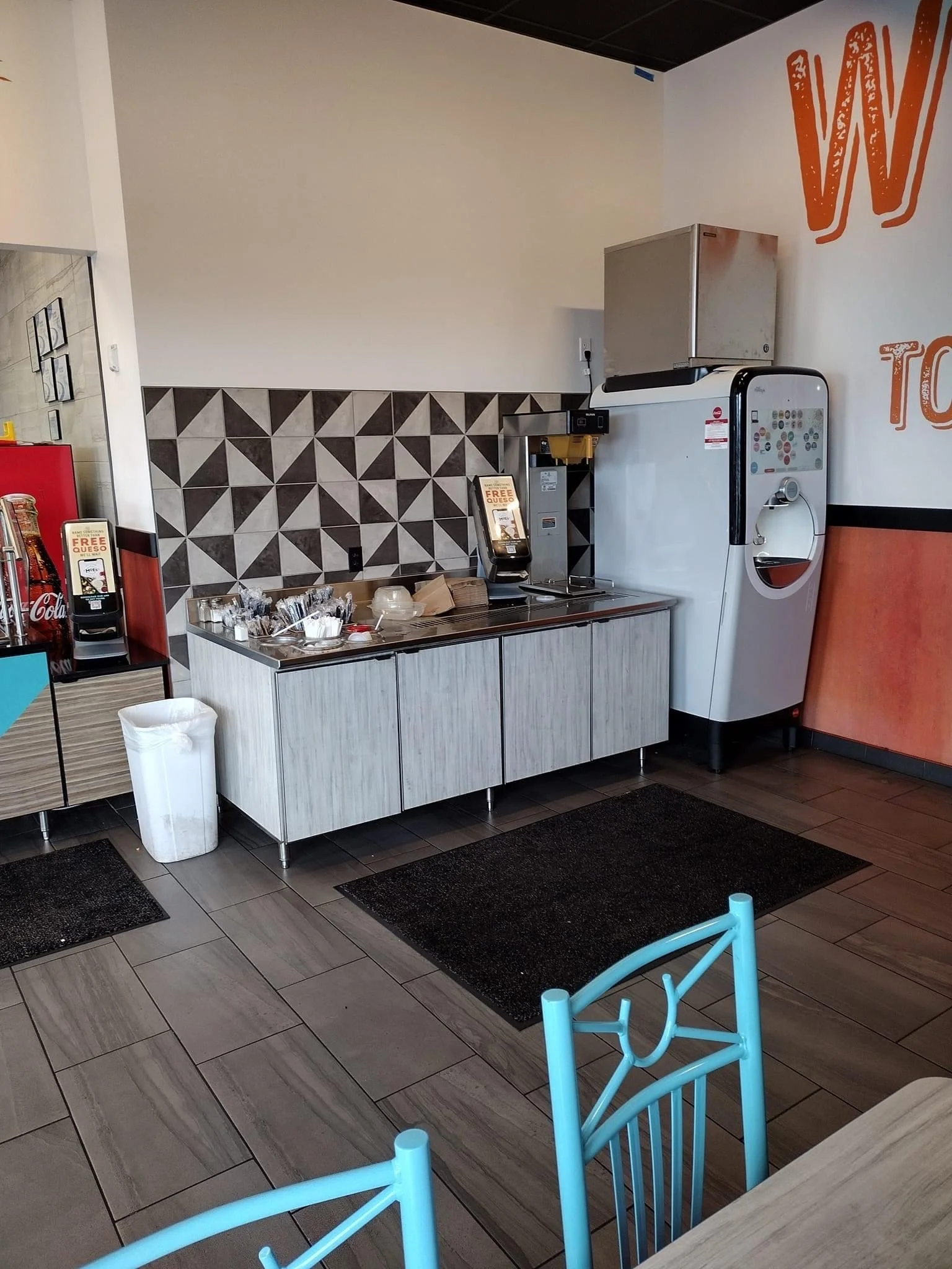 Restaurant beverage station with drink dispensers, condiment containers, and a geometric tile wall.