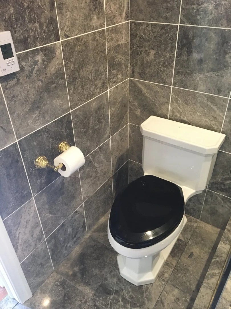 A bathroom with marble tiles featuring a white toilet with a black seat, a roll of toilet paper on a holder, and a wall-mounted thermostat.