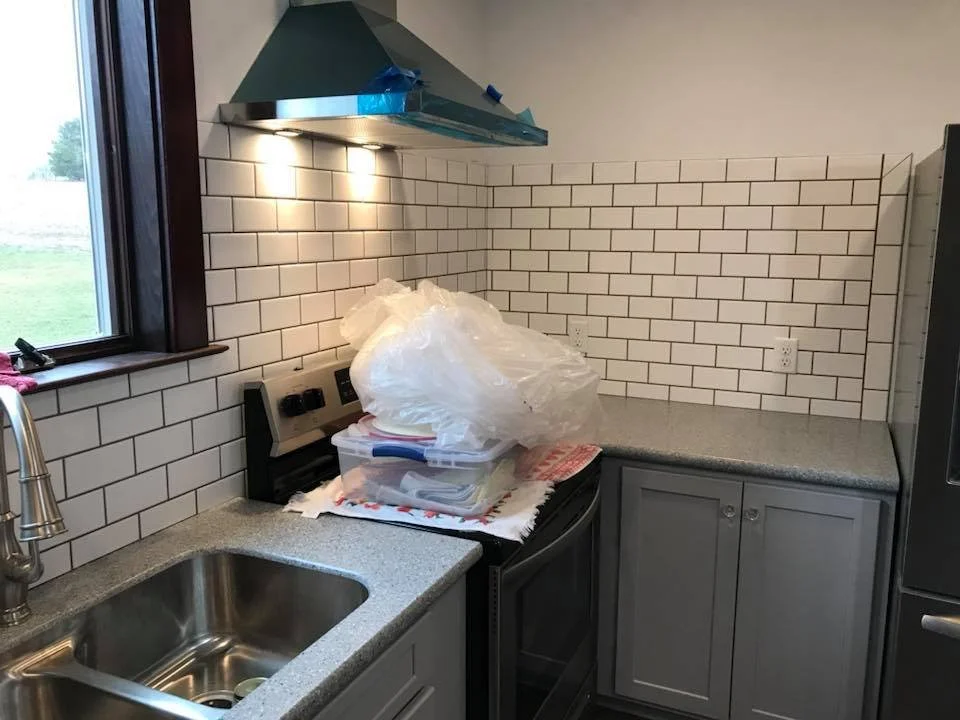 Modern kitchen corner with a stainless steel sink, white subway tile backsplash, and gray countertops. A range with a hood is installed, and plastic bags and containers are stacked on the counter. A window provides natural light.