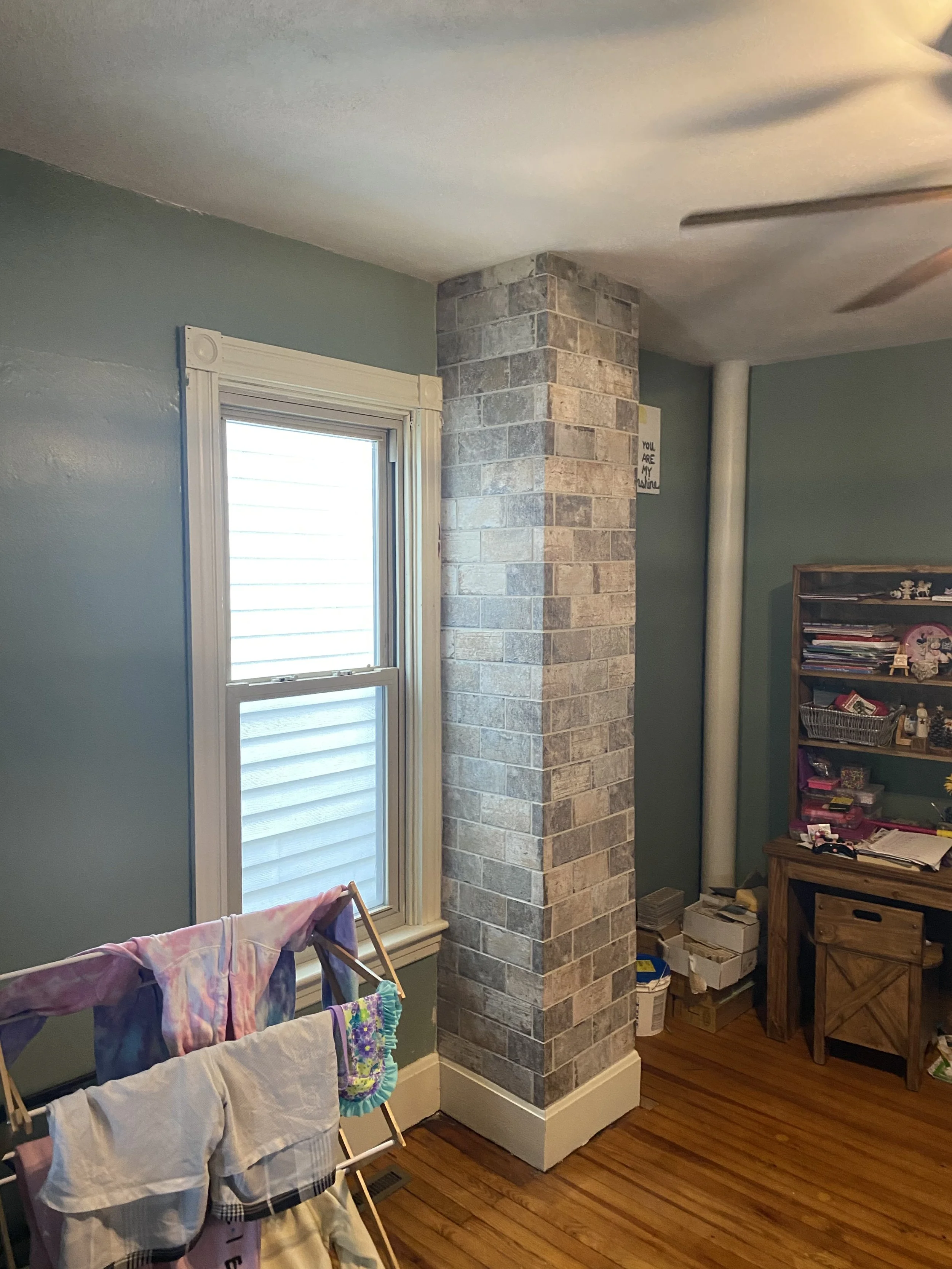 Room interior with a brick column, drying rack with clothes, window, and a wooden desk with shelves.