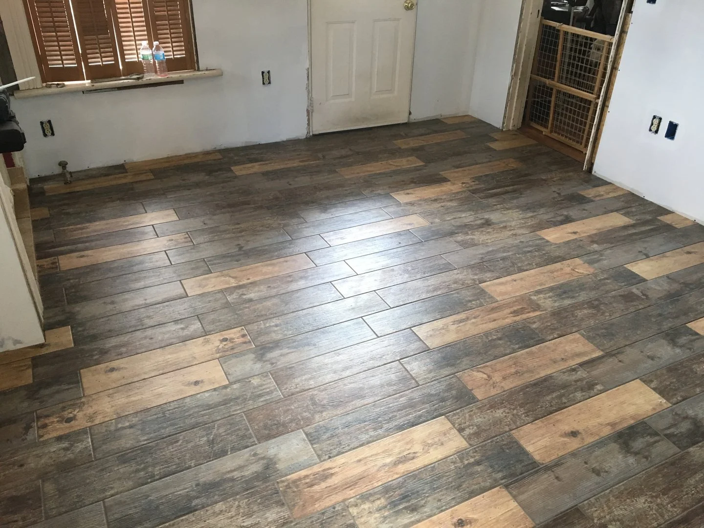 Room with wood-look tile flooring, white walls, a door, and window with shutters
