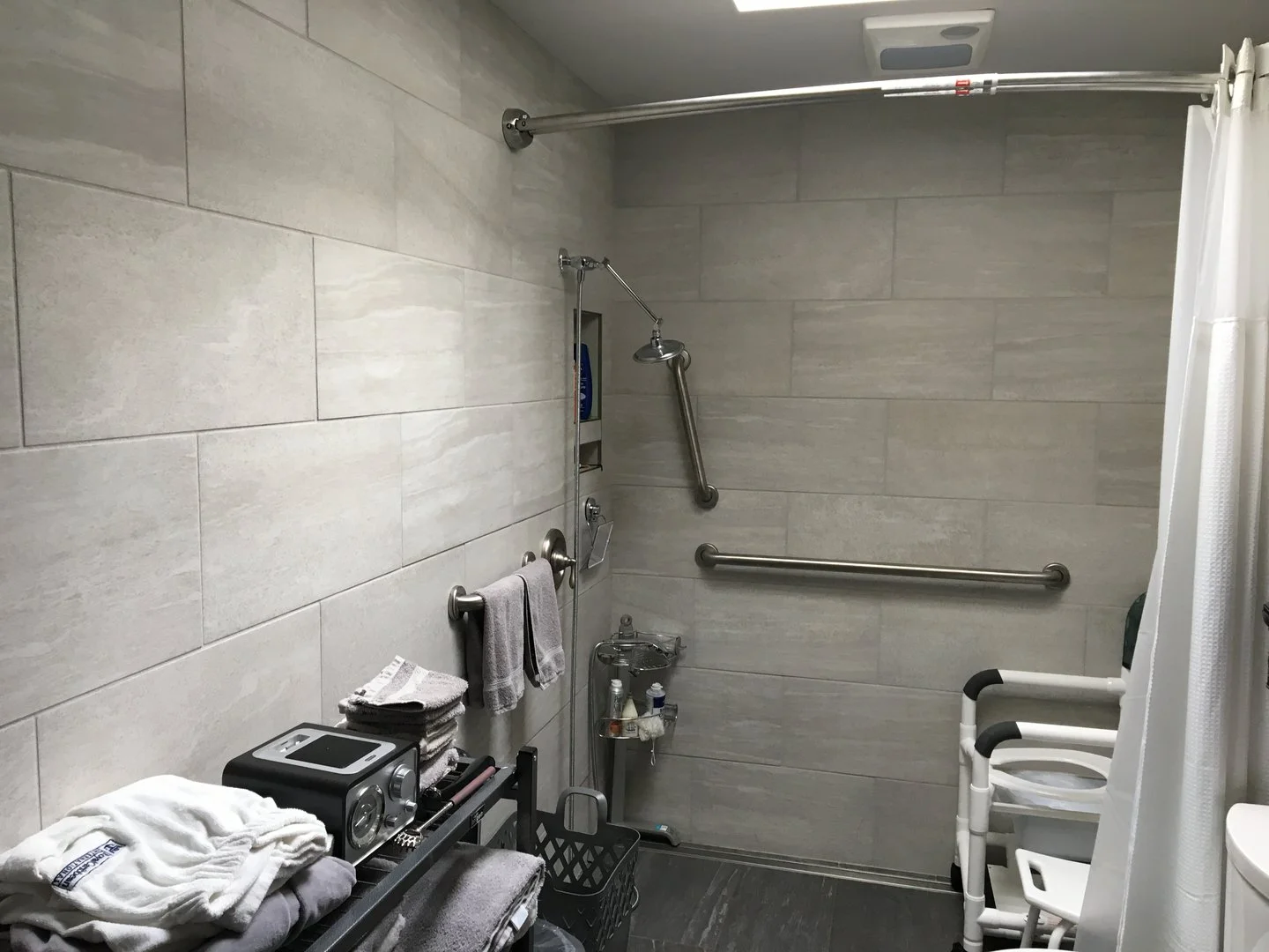 Accessible bathroom with tiled walls, shower area with grab bars, shower chair, and towels on a rack.